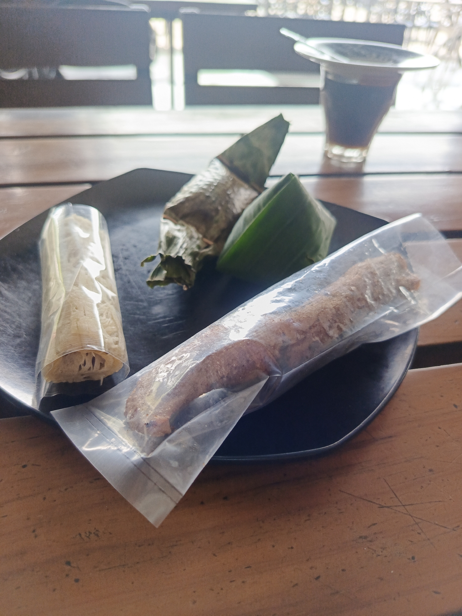 The image shows a black plate with three food items on a wooden table. One item is a cylindrical roll in clear plastic packaging, revealing what appears to be shredded or stringy material inside. Another item is wrapped in a leaf, and a third, longer item is also packaged in clear plastic, showing a dark brown substance with a slightly uneven texture. In the background, partially visible, is a clear glass cup containing a dark liquid, possibly coffee or tea, with a saucer and a spoon on top. Through a window behind the setup, a partially visible building with horizontal window slats can be seen. The location is in Bireuenmeunasahdayah, Indonesia.