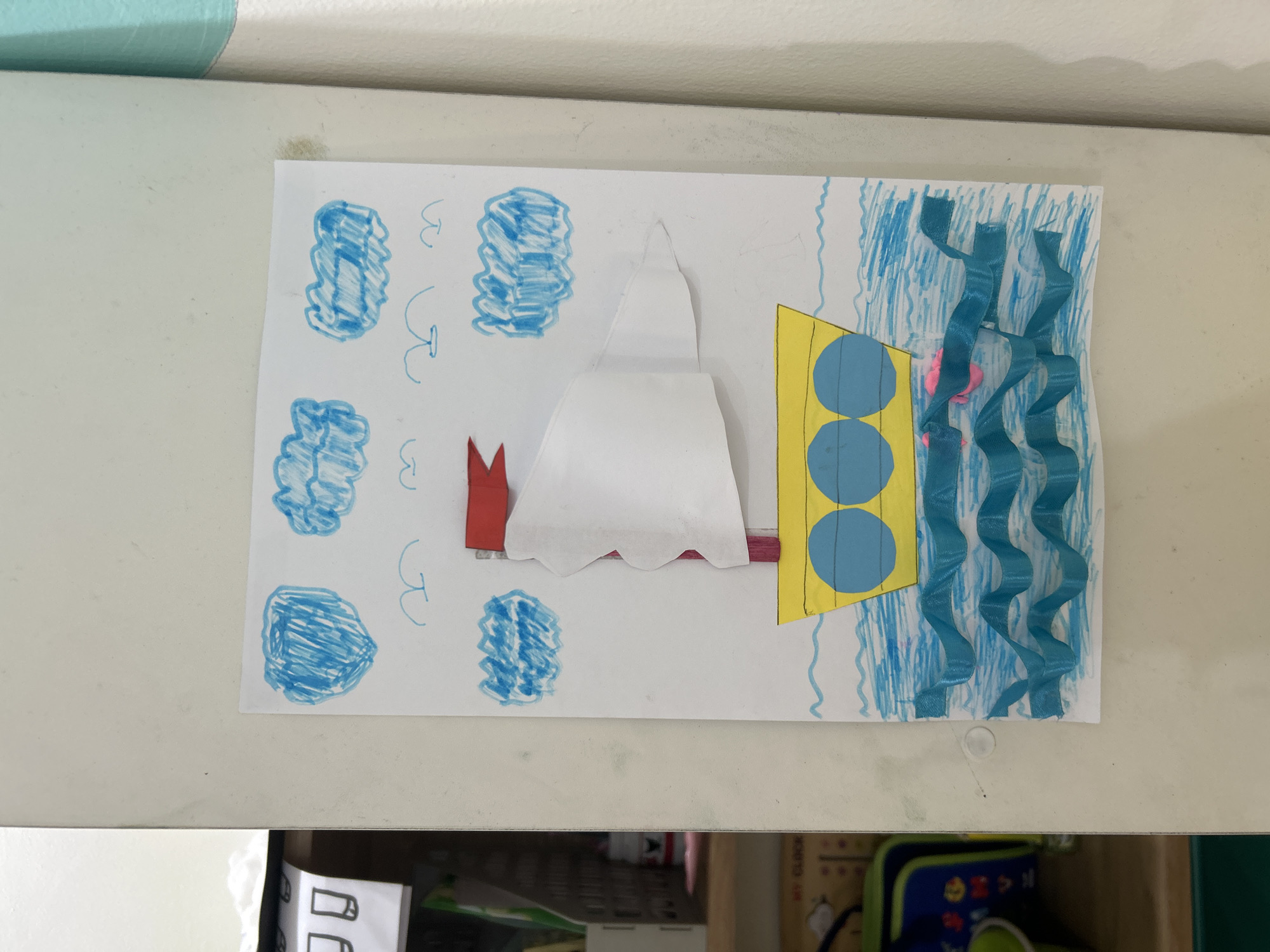A child's drawing of a yellow sailboat with blue circular windows is affixed to a white surface, possibly a refrigerator or wall. The sailboat has a red flag and a large white sail made of crumpled paper. The drawing depicts blue scribbled clouds in the sky and blue wavy lines for the water, with some three-dimensional blue ribbon-like material used to create textured waves at the bottom. The image is lit from the front. The background shows a slightly reflective surface on the left and a vertical light-colored curtain on the right. The context of Kuta, Indonesia, is not evident from the visual information provided in the image.
