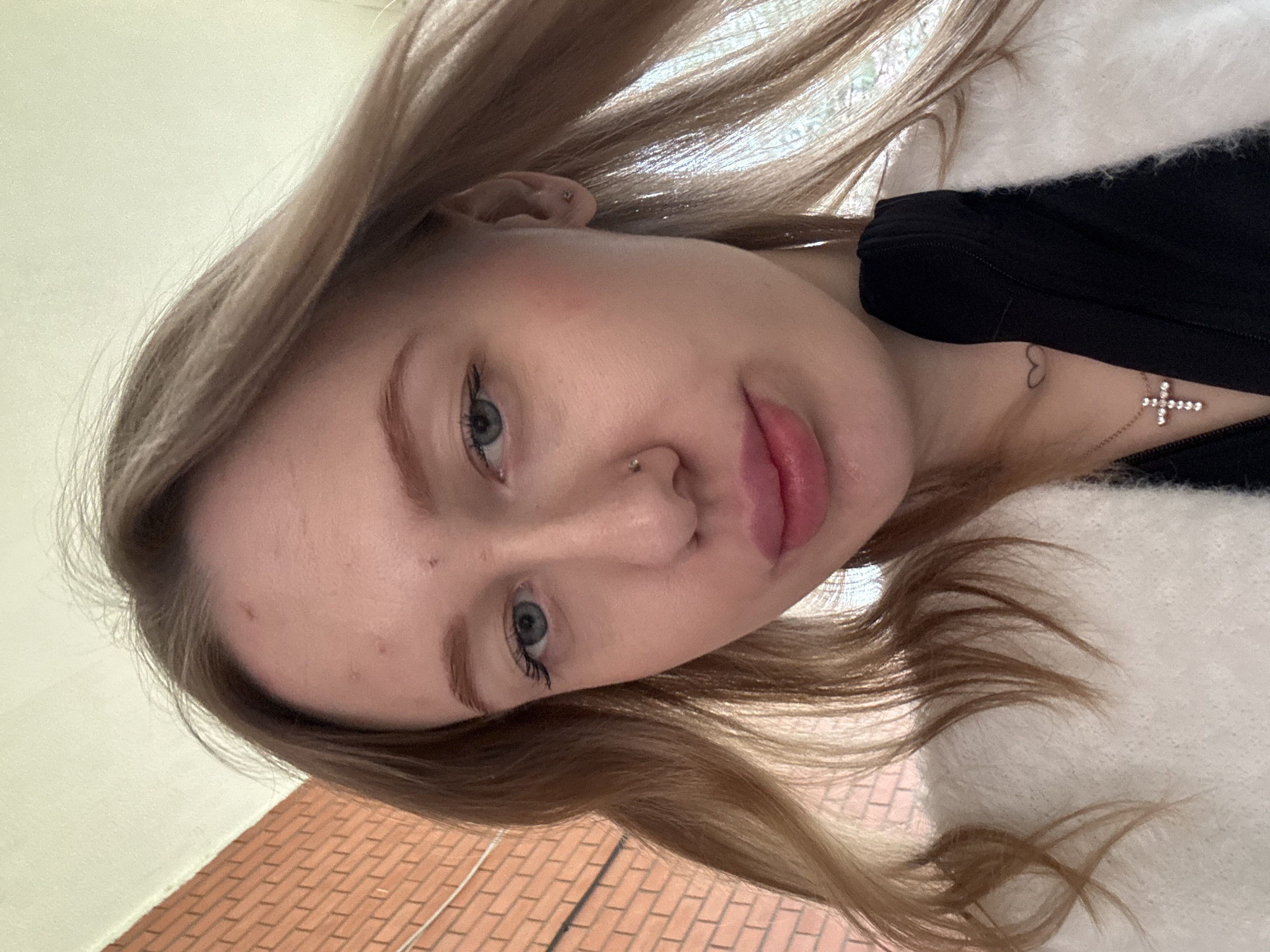 A medium, eye-level shot of a fair-skinned woman with light blonde hair, light eyebrows and blue eyes, who is wearing a black top and a white fluffy jacket. She has a nose ring in her left nostril. A small heart tattoo is visible on her chest near a necklace with a cross pendant. The background consists of a white wall, a brick floor, and a window. There is no indication that the picture was taken in Rostov-on-Don, Russia.