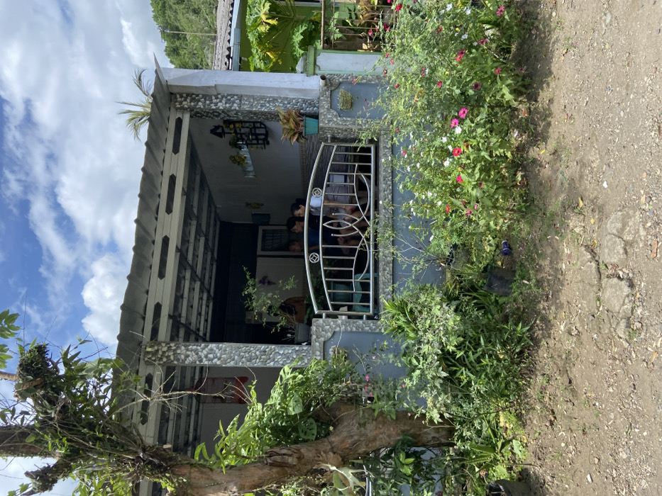 The scene is a residential area in Cipatat, Indonesia. The image shows a house with a tiled roof and a covered porch. The porch is enclosed by a black metal fence. There are three people sitting on the porch, two adults and one child. They are all looking towards the camera. The house is surrounded by a lush green garden. The plants in the garden are blooming, with pink and white flowers. The ground in front of the house is covered in gravel. The sky is blue and clear with a few fluffy white clouds. The scene is peaceful and tranquil.