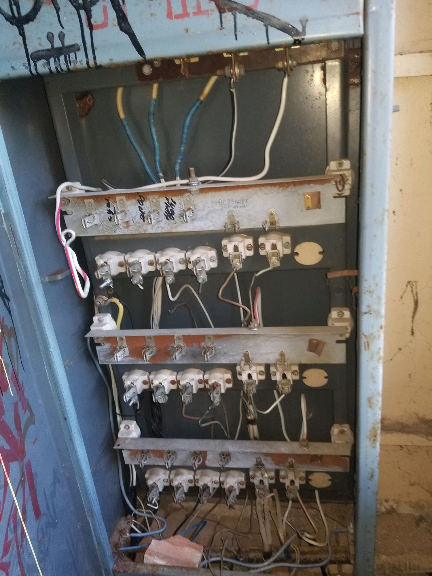 The image shows an interior view of an electrical panel or junction box. The panel is mounted on a wall and is made of metal. It contains multiple rows of electrical components, including fuses, wiring, and connectors. The panel appears to be old and weathered, with some rust and graffiti visible on the exterior. The lighting is dim, suggesting an indoor or dimly lit environment. The presence of graffiti suggests it may be located in an urban or semi-urban area.