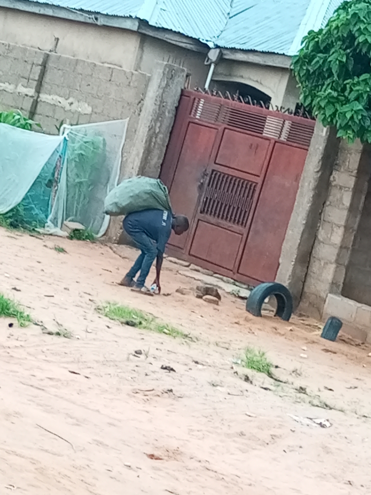 In Tempure, Nigeria, a man carries a large, green sack of goods over his shoulders, walking on a dirt path. He is wearing a dark blue shirt and pants, with brown sandals. The path is sandy with sparse vegetation and a few rocks. In the background, a red metal gate stands closed, flanked by a brick wall and a building with a light blue corrugated metal roof. To the left of the gate, a light-colored tent is visible, partially obscuring green plants. A discarded tire lies on the ground near the gate, and a smaller, dark object, possibly another piece of debris, is nearby.