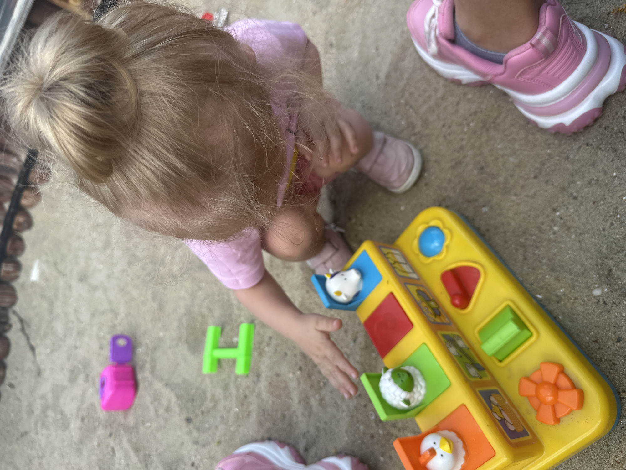 The image depicts a young child, likely a toddler, in a crouched position, playing with a yellow and blue pop-up toy featuring farm animals. The child has blonde hair styled in a bun and is wearing a pink t-shirt and pink shoes. Also visible are two pink and white sneakers, one on the child's left foot and another belonging to another person on the right side of the frame. Scattered on the sand-colored ground are a few other toys: a bright pink block and a smaller purple piece, and a green "H" shaped toy. The environment appears to be an outdoor area, possibly a backyard or playground, with sand or a similar granular surface. There is no visual information to place the scene in Donchovci, Bulgaria.