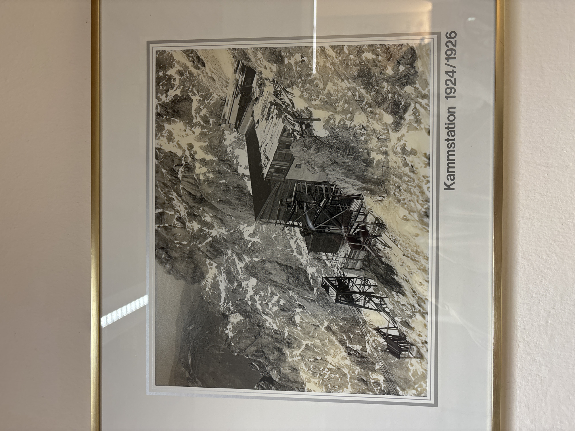 The photograph depicts a mountainous scene featuring a construction site, possibly for a cable car station, labeled "Kammstation 1924/1926". The structures are situated on a steep, rocky slope covered in patches of snow. Several wooden and metal scaffolding elements are visible, suggesting ongoing or recent construction. In the foreground and mid-ground, there are exposed rock faces and debris. The background shows a vast, rugged mountain landscape under a gray sky. The overall impression is one of a challenging, high-altitude construction project. The location is identified as being in Ehrwald, Austria.