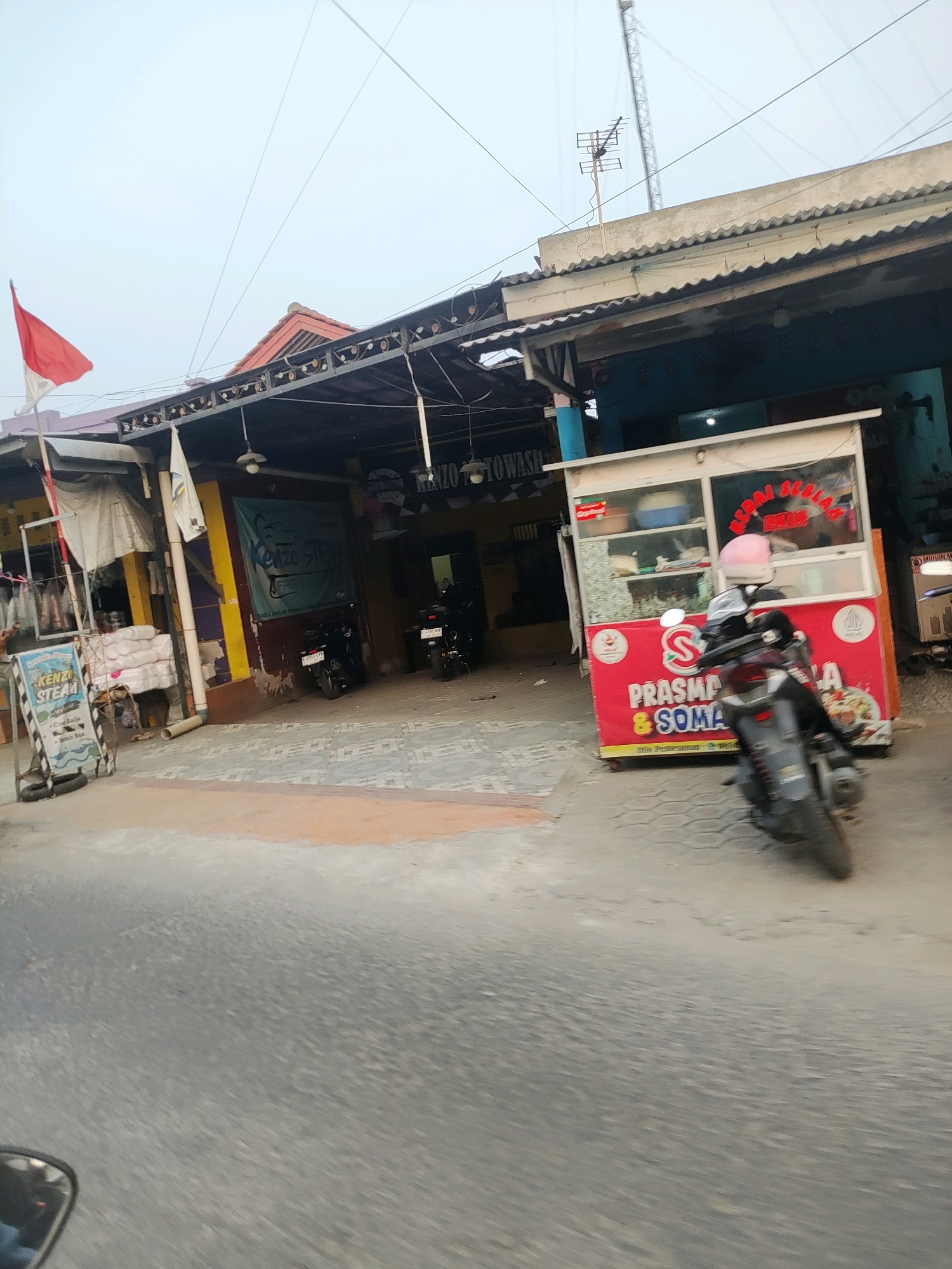 The image depicts a street scene in Wanajaya, Indonesia. A red and white Indonesian flag is visible on the left, attached to a pole extending from a building. In front of the building, there is a food cart with a red base and a glass display case. The cart has text in Indonesian, including "PRASMANA & SOMA." A motorcycle is parked in front of the food cart, and another motorcycle is parked in the background. The building appears to be a commercial establishment, with signs indicating services such as "KENZO STEAM." The ground is paved with concrete and asphalt. The sky is overcast.