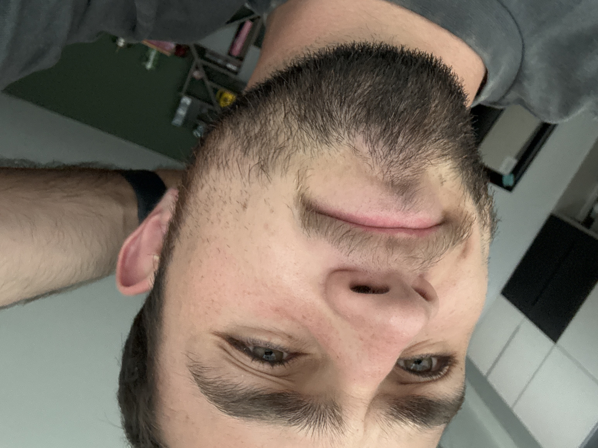 The image shows a close-up of a man's face and upper chest, taken from an upside-down perspective. He has a dark beard and short dark hair. He is wearing a grey shirt. His eyes are open. There is a dark-colored wristband on his left wrist. The background includes blurry objects such as shelving, and a wall with tile. There is no information to suggest this photo was taken in San Bernardino Tlaxcalancingo, Mexico.