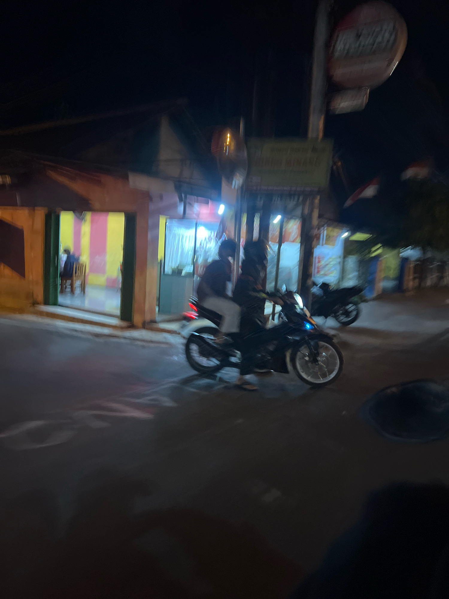 The image depicts a nighttime scene on a street in Magelang, Indonesia, likely near a small shop or restaurant. A motorcyclist, wearing a helmet and dark clothing, is stopped or moving slowly on a dark-colored motorcycle. The motorcycle has a silver rim on the front wheel and a red taillight. Another person, also wearing dark clothing, is seated behind the rider.
To the left of the motorcycle, a building with an open entrance is visible. Inside, a person is seated at a wooden chair. The interior walls are striped with yellow and pink. To the right of the building entrance, another section of the building has windows illuminated from within, with white curtains. A sign is mounted on a pole in front of the building, but its text is blurry and illegible. In the background, another motorcycle is parked. The street surface is dark and reflects some of the light. The overall atmosphere is dimly lit, with artificial light sources casting shadows and creating a sense of motion blur due to the low light conditions and likely movement of the camera or subjects.