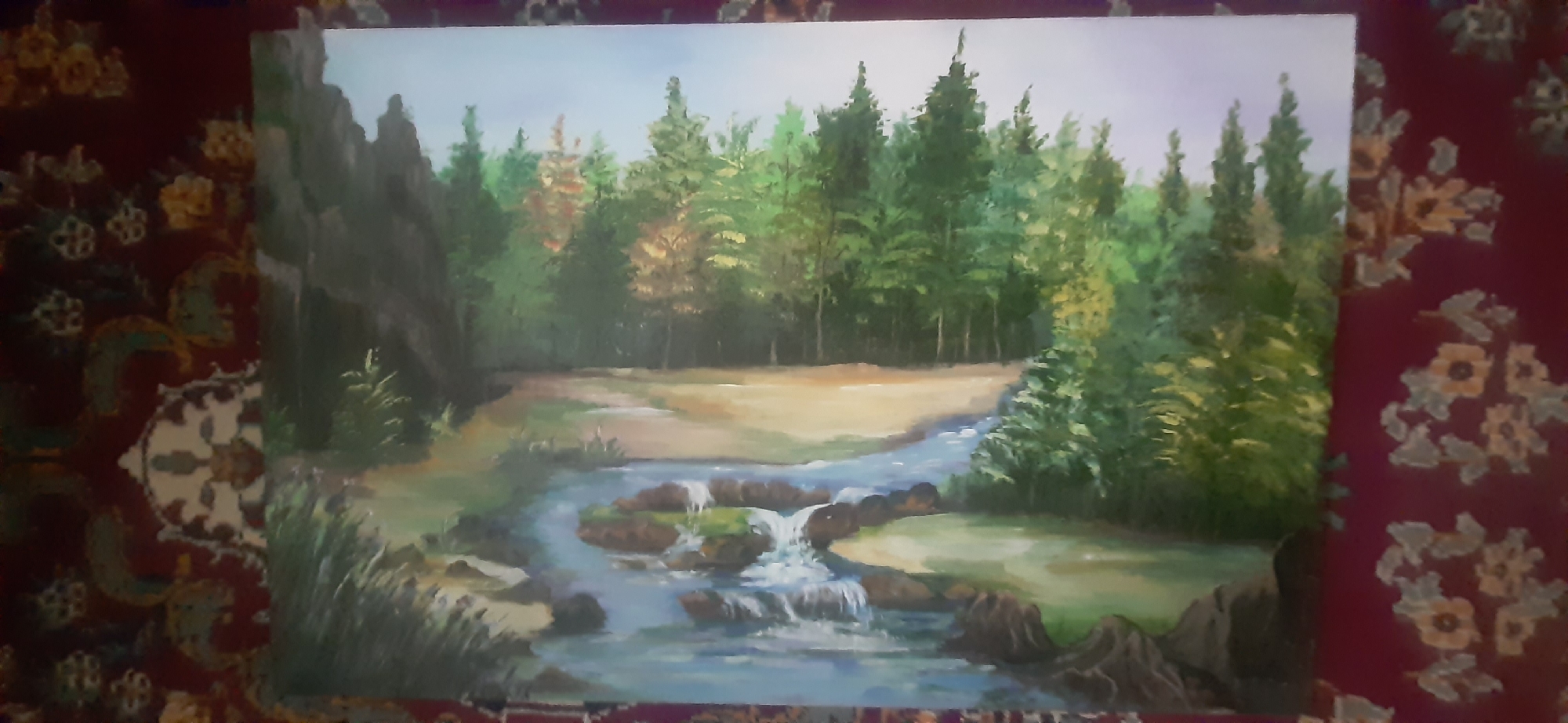 The image is a painting of a forest scene with a stream. The stream flows from the upper right to the lower left, with small waterfalls and rocks in the foreground. The stream is surrounded by green grass and trees. In the background, there is a dense forest with various shades of green and some patches of yellow and orange, suggesting autumn foliage. On the left side of the painting, there are large rocks. The sky is a light blue with some subtle color variation. The painting is displayed on a surface with a dark red, floral patterned fabric. The painting itself does not depict any part of the city of Yessentukskaya, Russia, or any specific geographic location. There are no people or animals visible in the painting. The scene is a natural landscape with a focus on trees and water.