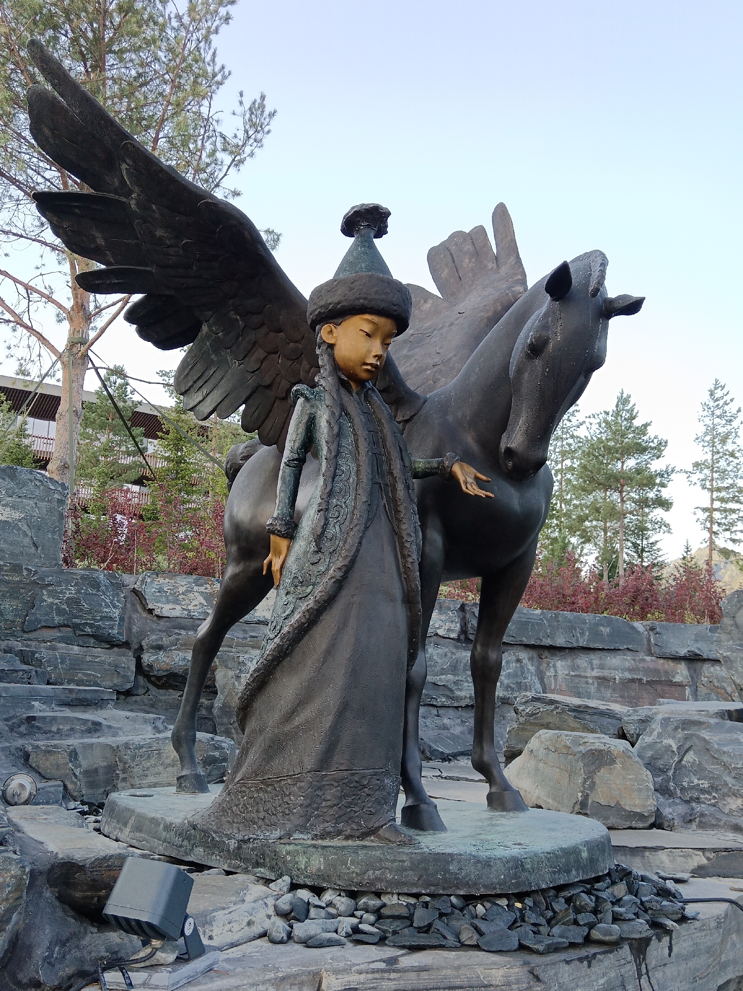 This is a photograph of a bronze sculpture depicting a young girl with large wings and a horse. The girl is wearing a traditional hat and a long, ornate coat. Her face is downcast, and she is reaching out towards the horse. The horse is standing beside her, with its head slightly lowered. The sculpture is situated outdoors, amidst a rocky landscape. In the background, trees and a building are visible. The overall setting suggests a park or outdoor exhibition space. The location is specified as the city of Ozyornoye (Maysky District), Russia. The lighting suggests it is daytime, possibly late afternoon or early morning, with soft, diffused light.