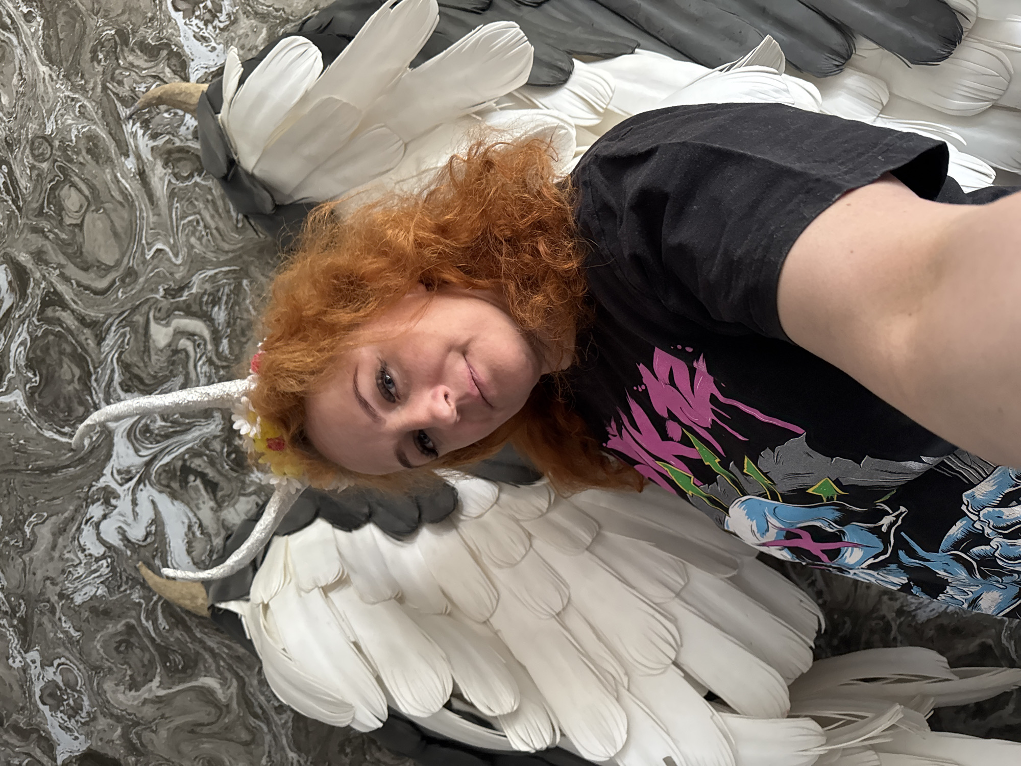 The image shows a woman with curly red hair, wearing a black t-shirt with a colorful graphic, lying on top of a sculpture of a swan. The swan has white and gray feathers and a white, curved neck and beak. The background is a marbled, swirled pattern of gray and white. The location is Chaikovsky, Russia.