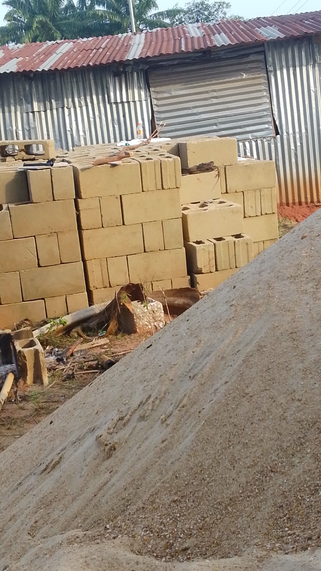 The image shows a construction site in Enugwu-Ukwu, Nigeria. A large pile of sand dominates the foreground, partially obscuring the view of stacked concrete blocks behind it. The blocks are arranged in columns, some with visible hollow cores. In the background, a corrugated metal structure with a red-roofed section is visible, indicating a temporary or informal building. Some vegetation is present.