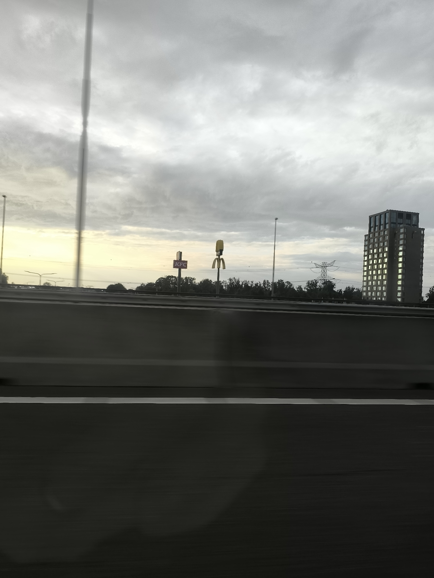 The image depicts a view from a highway in Lelystad, Netherlands, looking towards the horizon. On the right side of the frame, a modern, multi-story building with many illuminated windows is visible, bearing the word "HOTEL" vertically on its facade. To the left of the building, a large electricity pylon stands against a cloudy sky. Closer to the foreground, there are signs for fast-food restaurants, specifically a KFC sign and a McDonald's golden arches sign. The highway itself is prominent, with a concrete barrier in the midground and dark asphalt in the foreground, marked by a white line. The overall scene suggests a roadside view in an urban or semi-urban area, likely during daylight given the illumination of the building's windows.