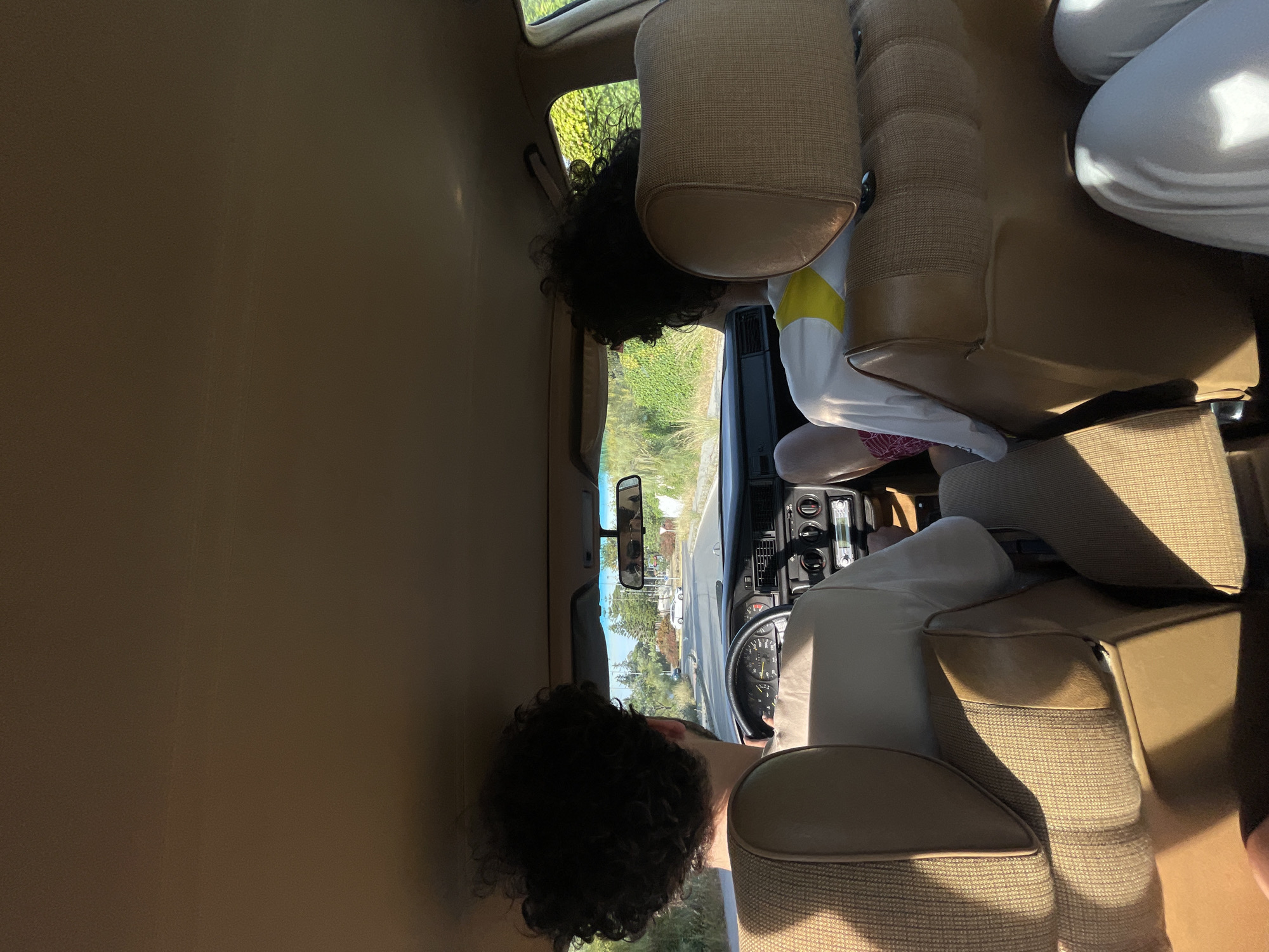 The interior of a vintage car is shown, with two people present. One person is behind the steering wheel, apparently driving, while another individual sits in the back passenger seat. The car is situated on a road, and greenery is visible through the windows, suggesting an outdoor location. Based on the metadata, the location is El Puerto de Santa María, Spain. Details like the car's dashboard, seats, and the clothing of the occupants are also visible.