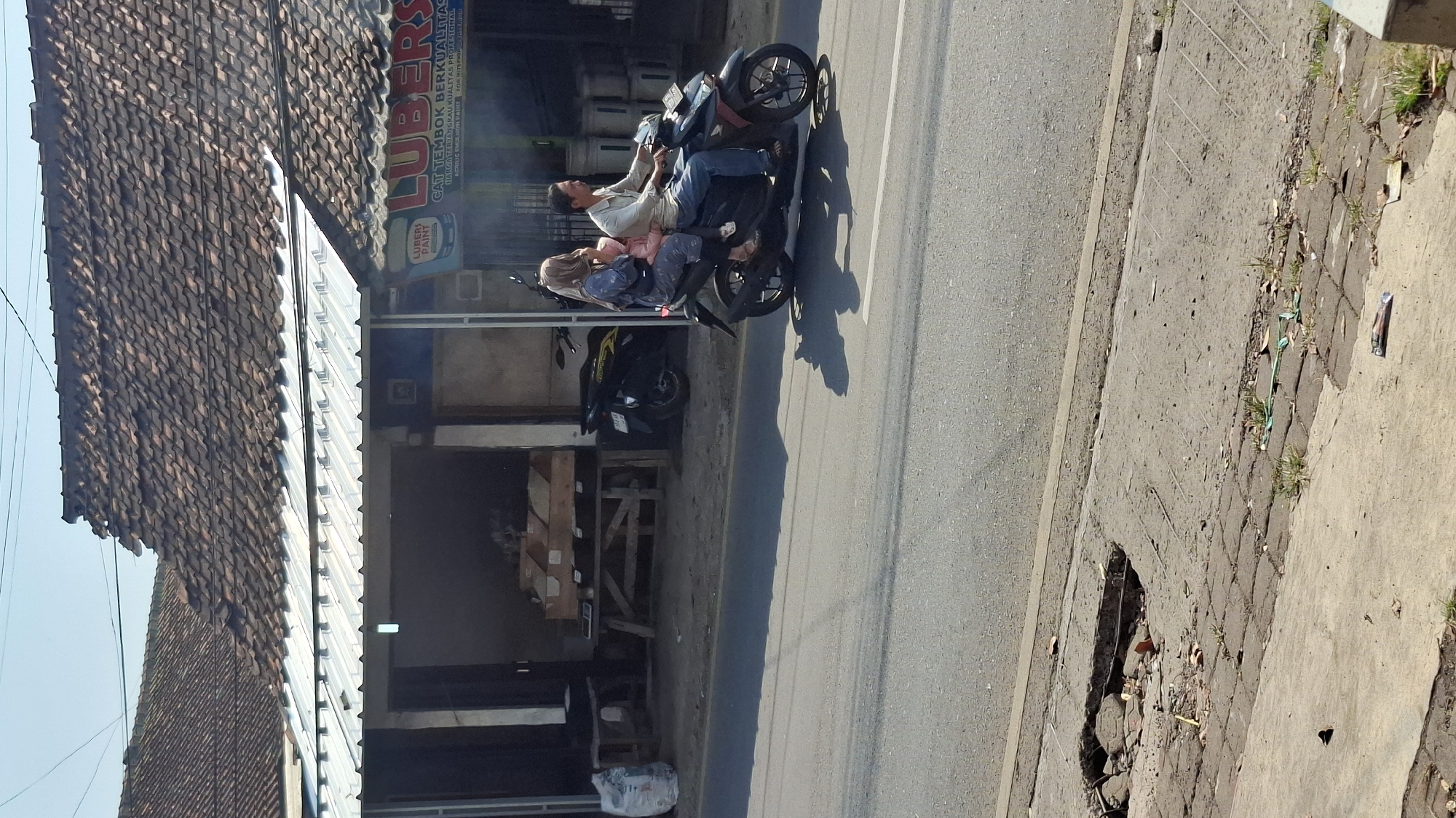 In the city of Cilaku, Indonesia, on a bright, sunny day, a family of three rides a black motorcycle along an asphalt road. The father, wearing a light-colored shirt and blue jeans, drives the motorcycle. Next to him sits a woman wearing a patterned headscarf and a light blue long-sleeved shirt, holding a small child in a pink jacket. The child is facing forward. The motorcycle casts a shadow on the road.
In the background, a building with a red tile roof and a metal awning is visible. Several motorcycles are parked in front of the building. Signs on the building indicate it is a store selling "LUBERS PAINT." The building appears to be a commercial establishment, possibly a paint store or hardware store.
In the foreground, the edge of the road is lined with a paved sidewalk. A section of the sidewalk is broken, revealing exposed concrete and rebar. Debris, including small stones and bits of plastic, is scattered around the broken pavement. The overall scene depicts a typical street view in a developing urban or suburban area.