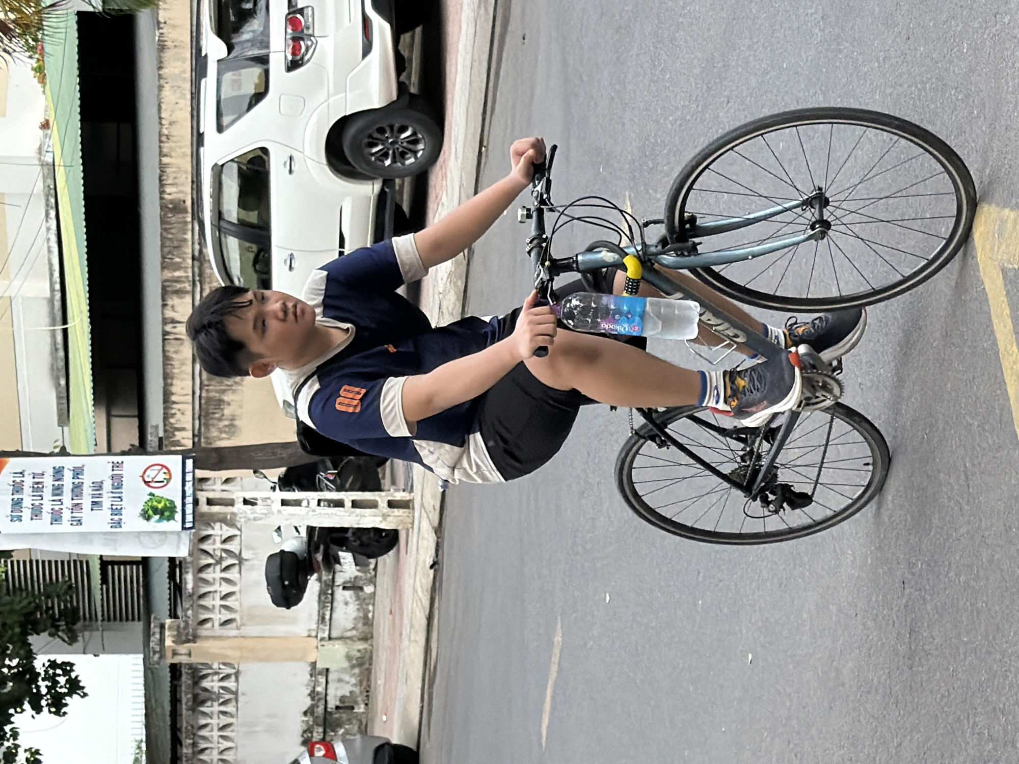 A young male is riding a bicycle on a paved street in Nha Trang, Vietnam. The boy is wearing a navy blue and white short-sleeved t-shirt with the number "00" on the sleeve, black shorts, and athletic shoes. A water bottle is attached to the frame of his blue bicycle, which has black wheels. In the background, a white SUV is parked on the side of the road. A sign with Vietnamese text and two circular icons, one with a green vegetable and a prohibition symbol, and another with a cigarette and a prohibition symbol, is visible on a pole to the left of the boy. The street appears to be lined with buildings and vegetation.