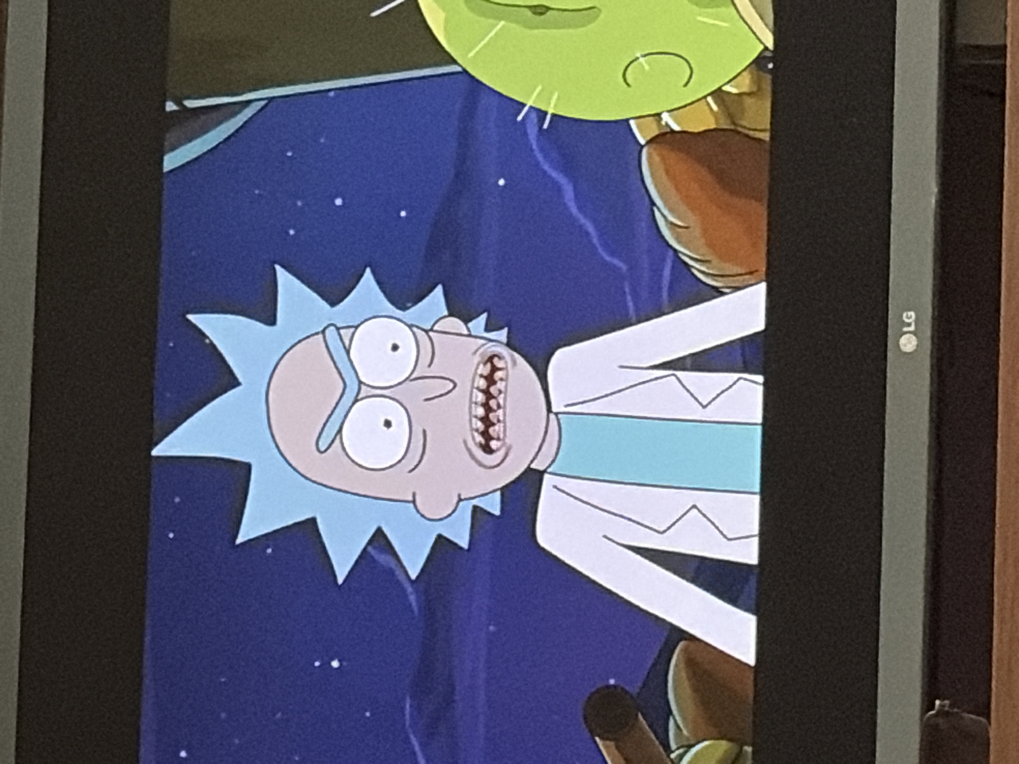 An animated character, Rick Sanchez, with spiky blue hair, a lab coat, and a panicked expression, is depicted in the foreground. He is looking to his left with wide, staring eyes and a grimace revealing his teeth. Behind him, a green, spherical character with a single eye and a curved line for a mouth is visible on the right. The background is a dark blue night sky with scattered white dots representing stars. Rick is wearing a light blue shirt under his white lab coat, which is open in the front. The scene appears to be a still from an animated television show, displayed on a television screen with an "LG" logo at the bottom.