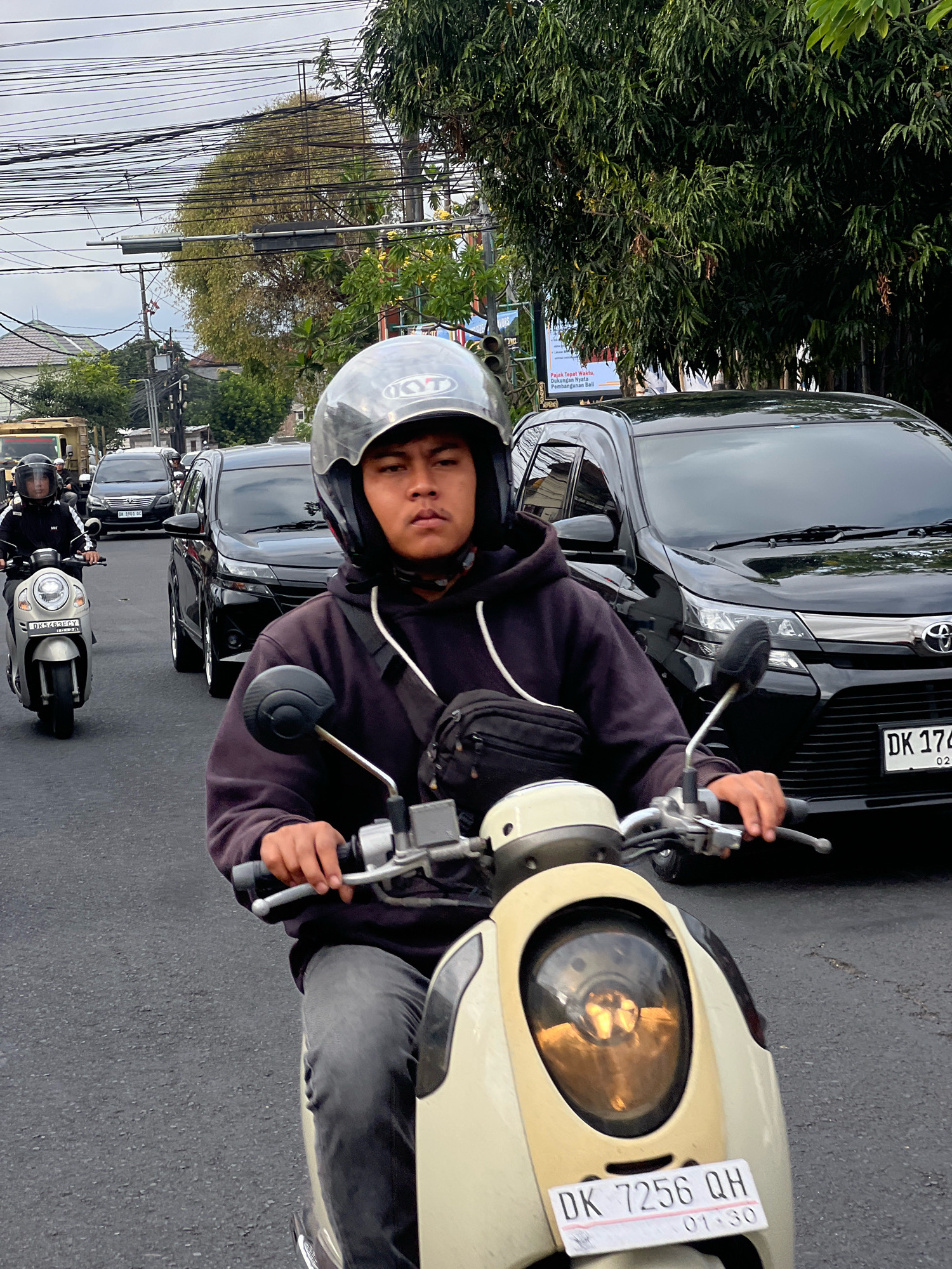 A young man wearing a black hoodie and a silver helmet is riding a cream-colored scooter on a paved street in Denpasar, Indonesia. He is looking forward, holding the handlebars. Another person is riding a silver scooter to his left, also wearing a helmet. Several black cars are in the background, including a Toyota Avanza. Overhead, there are numerous electrical wires and poles. Trees line the right side of the street. The sky is overcast. The license plate of the scooter in the foreground reads "DK 7256 QH".