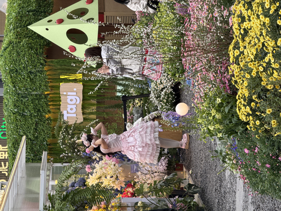 The scene depicts a vibrant outdoor garden installation within an urban setting in Liangjiang, China, likely a commercial or public display area. In the foreground, numerous flowering plants are arranged in beds, featuring prominent yellow chrysanthemums, pink daisies, and various blue and white blooms. A dark gravel path runs through the left side of the frame, leading to a circular, glowing white light fixture positioned amidst the foliage.
Two individuals are the primary subjects. On the left, a person with dark hair, adorned with a pink floral headband and wrist cuffs, wears a layered pink Lolita-style dress with white lace trim, white tights, and pink platform shoes. This individual stands in profile, facing left, with hands raised near the head in a posed manner. To the right and slightly behind, another person, with dark hair, is seen from the back, wearing a light grey jacket over a colorful patterned skirt. This individual holds a smartphone in both hands, extended forward, appearing to photograph the person in the pink dress or the surrounding floral display. A third person is partially visible on the far right.
The background features a building facade covered extensively with green foliage, creating a vertical garden effect. A prominent light brown wooden sign with the white text "Tagi." is mounted on this green wall. To the right of the sign, a large, light green, triangular pyramid-shaped structure is visible, adorned with red apple details and circular cutouts. Overhead, partial glimpses of a structure with "City" text are visible. The overall environment is brightly illuminated, suggesting daytime or an exceptionally well-lit evening event.