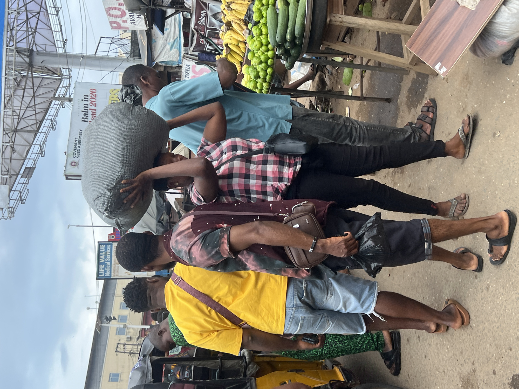 The image depicts a busy street scene in Nkpor, Nigeria, likely a market area. In the foreground, several individuals are visible. A woman is carrying a large, gray, rough-textured sack on her head, dressed in a pink and black plaid shirt and black pants. To her right, a man in a light blue t-shirt and gray pants stands. To the left of the woman, a man in a yellow t-shirt and ripped denim shorts is positioned. Another man, wearing a maroon and tie-dye patterned shirt and dark pants, is in the center, holding a black plastic bag. A person wearing a green patterned garment and black sandals is partially visible on the far left, next to a yellow auto-rickshaw. The background reveals market stalls with produce, including rows of green fruits and bananas. Several signs are visible, including one reading "LIFE VALUE Medical Services" and another for "COVENANT SEED ASSEMBLY" with text mentioning "Balm in Gilead 2026". The overall environment suggests a bustling commercial district with pedestrian and vehicular traffic.