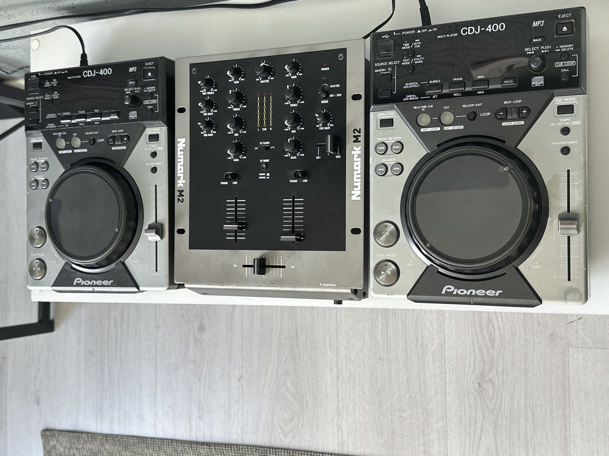 The image shows a DJ setup consisting of two Pioneer CDJ-400 CD players and a Numark M2 mixer. The equipment is arranged on a white surface, possibly a table or shelf. The CD players are positioned on either side of the mixer. The Numark M2 mixer is centrally located between the two CDJ-400 players.
The left CDJ-400 player displays a control panel with buttons, knobs, and a jog wheel. The front panel prominently features the "Pioneer" logo.
The Numark M2 mixer has multiple knobs, faders, and switches. The brand "Numark M2" is visible on the side of the mixer.
The right CDJ-400 player also shows a similar control panel to the left, with the "Pioneer" logo displayed on the front panel. Cables are visible connecting the equipment.
The background is blurred, showing a light-colored floor or surface. The image is taken from a slightly elevated perspective.
There is no location information in the image. No people, subjects, or actions are shown in the image. The objects are music DJ equipment. The environment is an interior surface, possibly a home or studio.