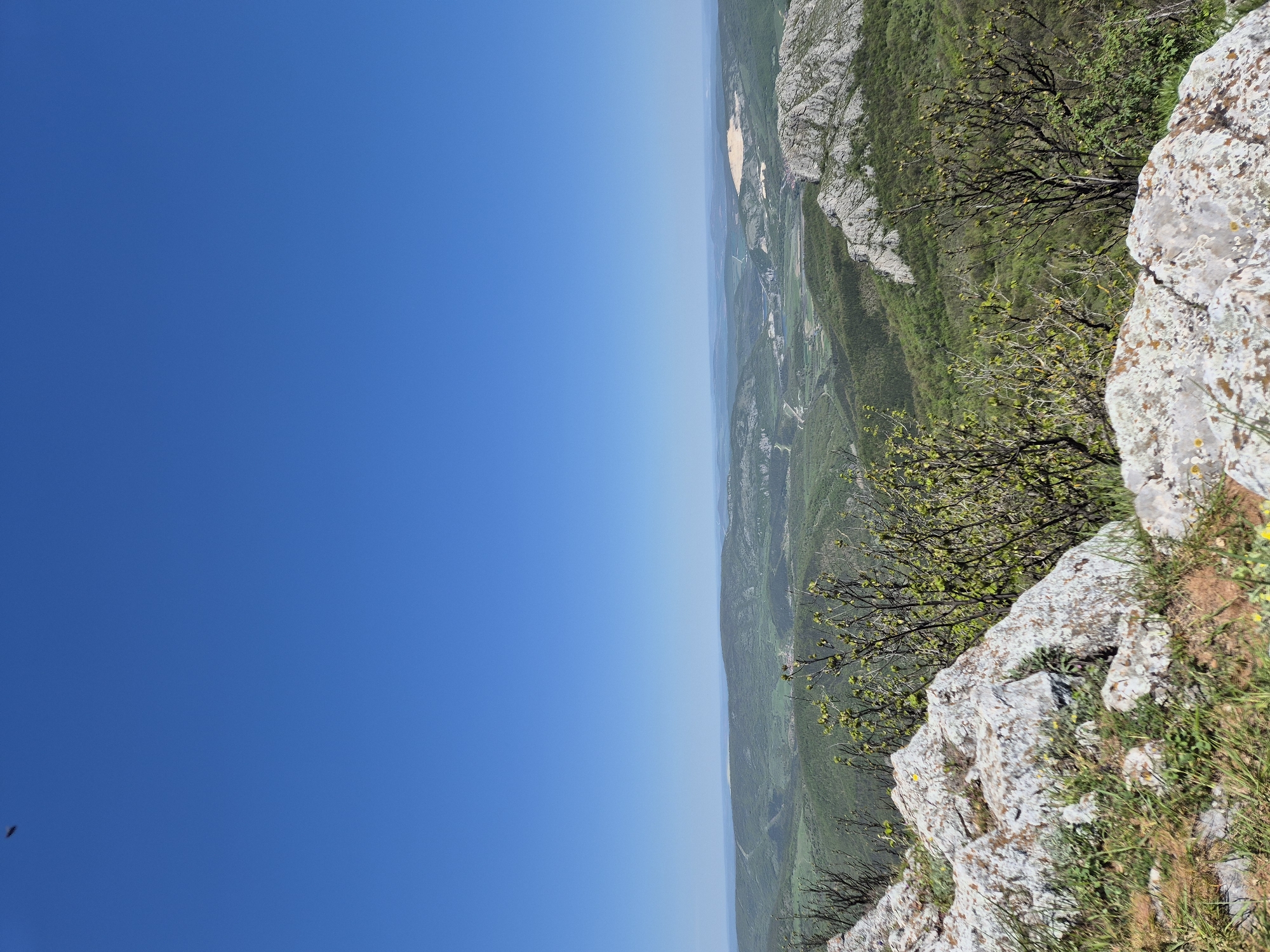 The image depicts a landscape view from what appears to be a high point, likely a cliff or mountain edge, near Bilka, Bulgaria. The right side of the image shows rocky terrain, vegetation, including trees and grass, growing on the cliffside. The left side of the image is dominated by a clear blue sky. In the middle distance, there is a view of the landscape below, which includes green valleys and possibly some bodies of water, blending into a hazy horizon. The overall impression is a scenic, elevated view on a clear day.