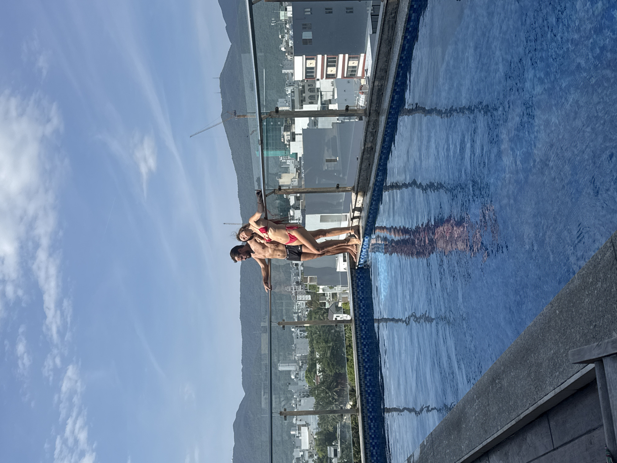 A man and a woman are posing on the edge of an infinity pool that overlooks the city of Đà Nẵng, Vietnam, and the sea. The man is standing with his feet on the edge of the pool and has one hand on the glass barrier behind them. The woman is straddling the man while holding onto his shoulders. The man is wearing black swimming trunks. The woman is wearing a red bikini. The pool is surrounded by a glass barrier. The background includes the city, mountains, and the sky. The sky is blue with scattered clouds. Reflections of the man and woman are visible on the surface of the water.