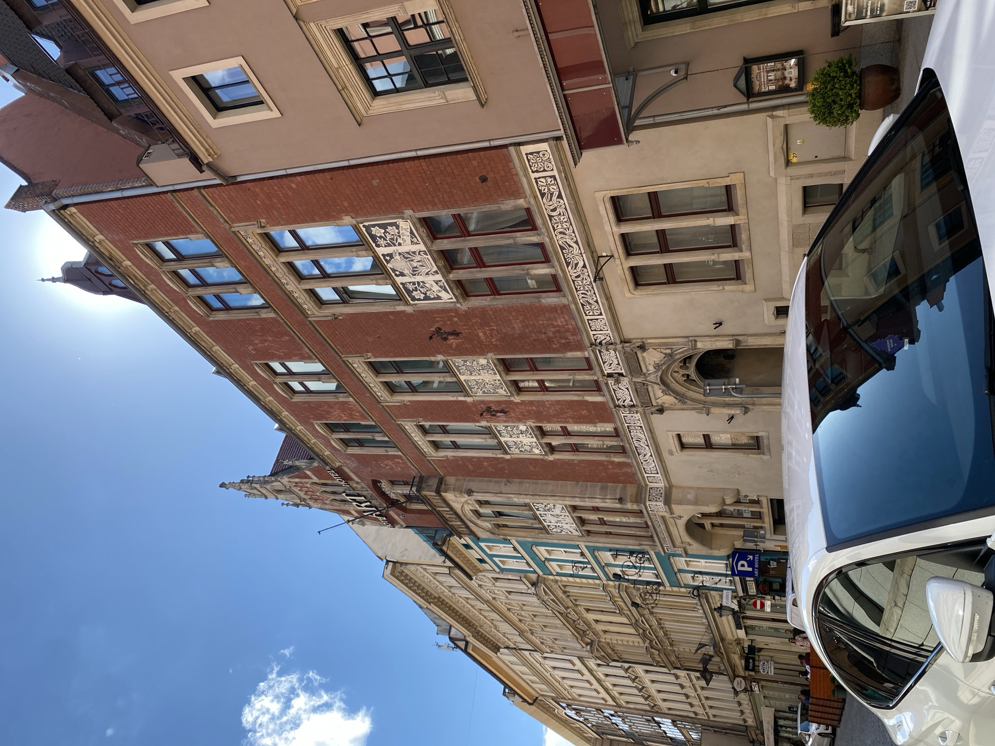 This detailed image captures a street scene in Wrocław, Poland, under bright, clear daylight.
In the foreground, the hood, windshield, and a portion of the driver's side front of a white four-door sedan are prominently displayed, reflecting the surrounding buildings and the blue sky.
The midground features a narrow urban street lined with multi-story buildings. On the left, a row of predominantly cream or light beige buildings extends into the distance, characterized by ornate architectural details, numerous windows with dark frames, and some balconies. One building further down the street exhibits a distinct turquoise-colored section on its upper floors. Several businesses operate at street level, with outdoor seating areas enclosed by wooden barriers, and signs such as "TULIP" and "ANIXER" are partially visible.
On the right side of the street, the most prominent structure is the "ART HOTEL," a multi-story building with a reddish-brown brick facade. It features dark-framed windows and decorative lighter-colored panels between floor levels, adorned with intricate white and dark grey patterns and stylized imagery. The name "ART HOTEL" is inscribed in dark letters on a lighter background near the ornate gable roofline. An elaborate arched entrance is visible at street level. Immediately to its right is another multi-story building, lighter beige in color, with simpler dark-framed windows and a dark-red canopy over an entrance. A potted green bush is situated near this entrance. A blue parking sign indicating "P ART HOTEL" is affixed to a pole or the adjacent building.
The street itself appears to be paved with cobblestones or patterned pavers. In the mid-distance, several individuals are visible, either walking on the pavement or seated at outdoor tables.
The background consists of a bright blue sky with a few scattered white cumulus clouds in the upper left quadrant. Sunlight is strong, casting some glare in the upper right portion of the frame.