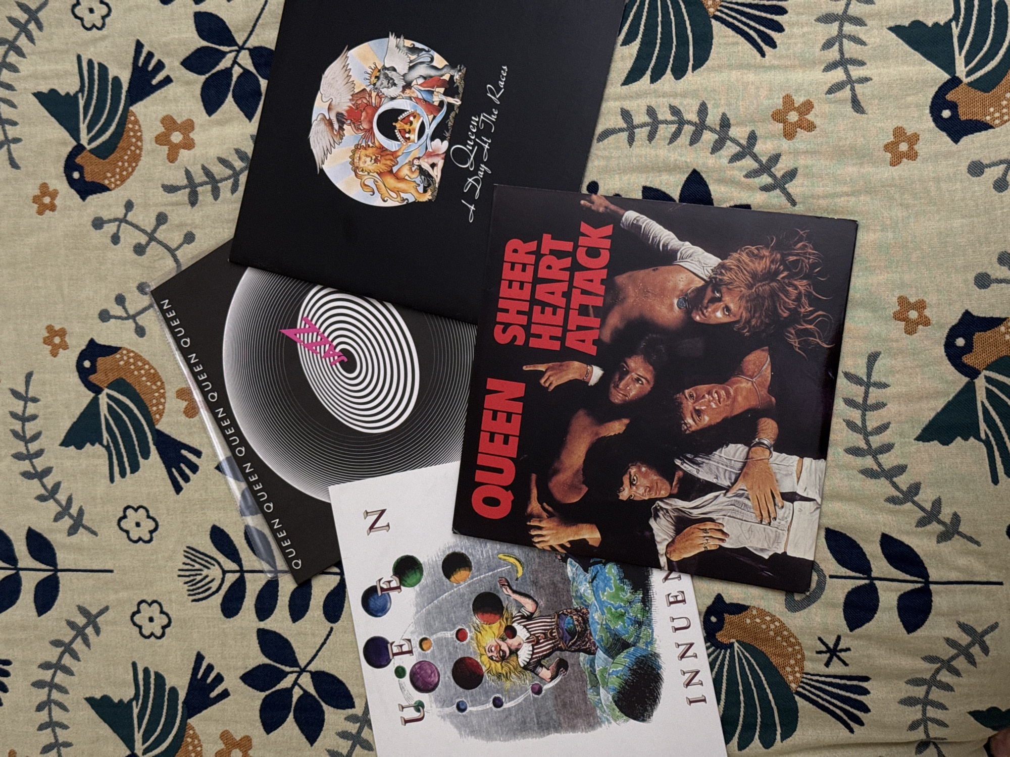 The image shows a close-up, overhead shot of four Queen vinyl records arranged on a patterned fabric. The album covers visible are "A Day at The Races", "Sheer Heart Attack", "Innuendo", and one with a black-and-white swirl design, potentially "Jazz". The fabric has a beige background with a repeating pattern of dark blue and dark green birds and stylized floral elements. The scene does not appear to be located in Moscow, Russia.