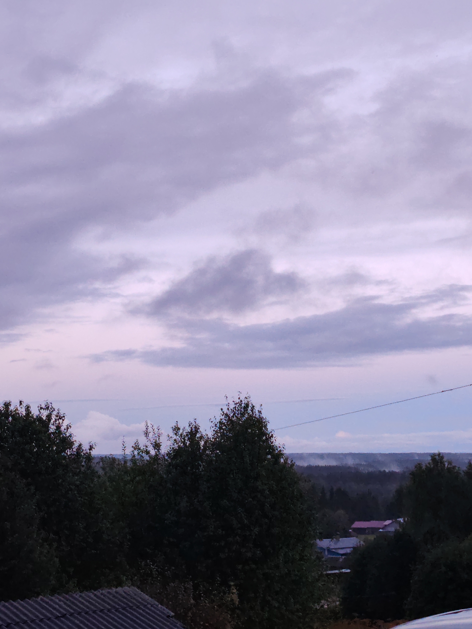 The image depicts an overcast sky over a rural landscape. The sky is filled with diffuse, lavender-hued clouds, with some areas appearing lighter than others, suggesting a twilight or pre-dawn setting. In the foreground, the dark silhouettes of dense trees are visible, their leaves and branches obscuring the view of the ground. A corrugated metal roof, likely part of a building, occupies the lower left portion of the frame. Beyond the trees, a distant village or settlement can be seen, with several houses featuring red and blue roofs. A thin, dark horizontal line, possibly a power line, stretches across the middle ground. The background consists of a hazy, forested horizon, with faint indications of mist or low-lying clouds. The overall impression is one of a quiet, rural evening or morning in Чухлэм, Russia.