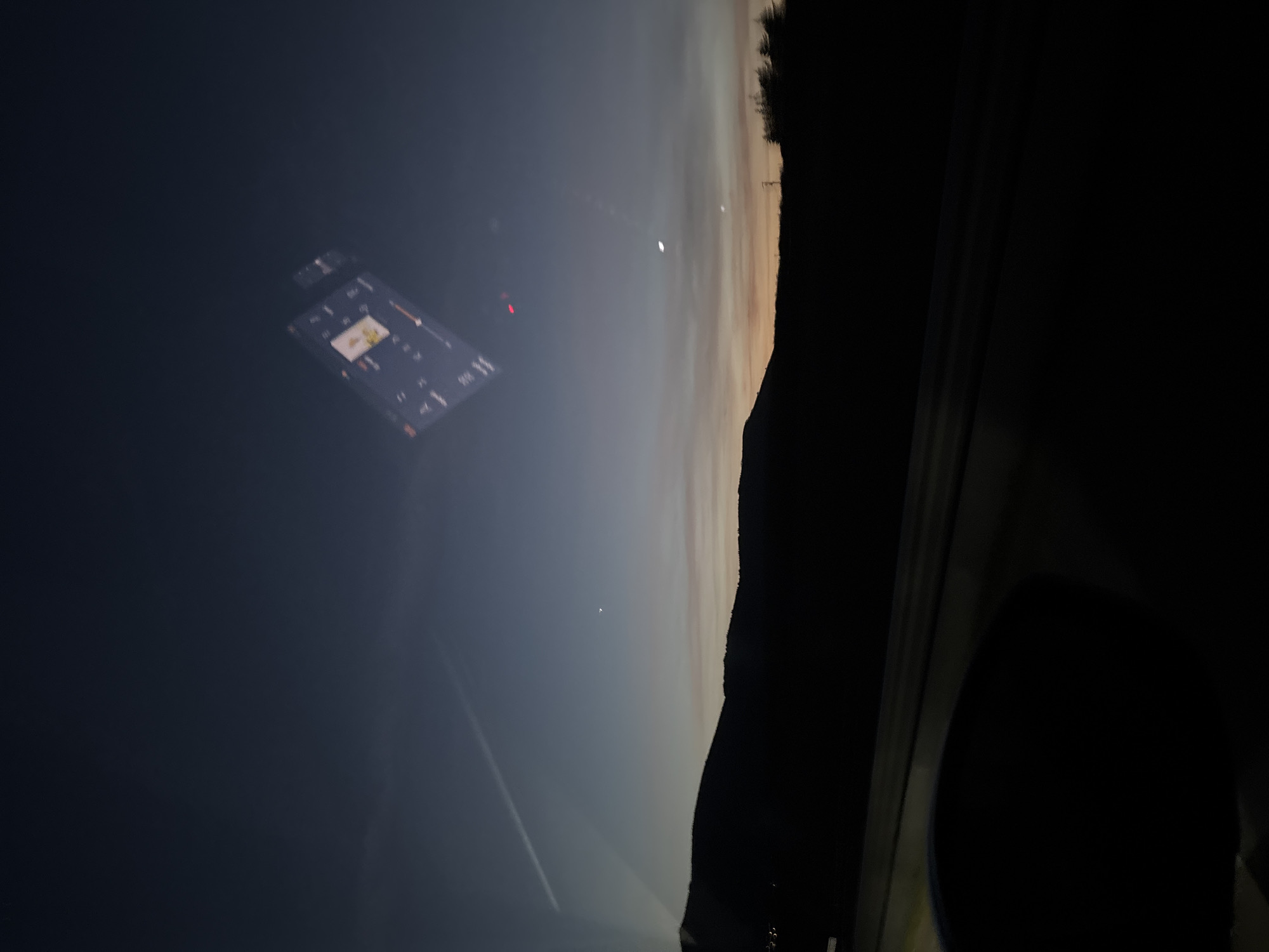 The image, rotated counterclockwise, depicts a night scene with a view from inside a vehicle. A smartphone or similar device is visible on the dashboard, displaying an interface with buttons and graphics. Outside the vehicle, the horizon shows a transition from twilight to darkness, marked by the silhouette of a land feature, possibly a hill or mountain range. Above the horizon, a bright celestial object (likely a planet) and the moon are visible. A contrail from an aircraft is observed in the sky.
Without further information, pinpointing the exact location in Mirandela, Portugal is not possible from visual data alone.