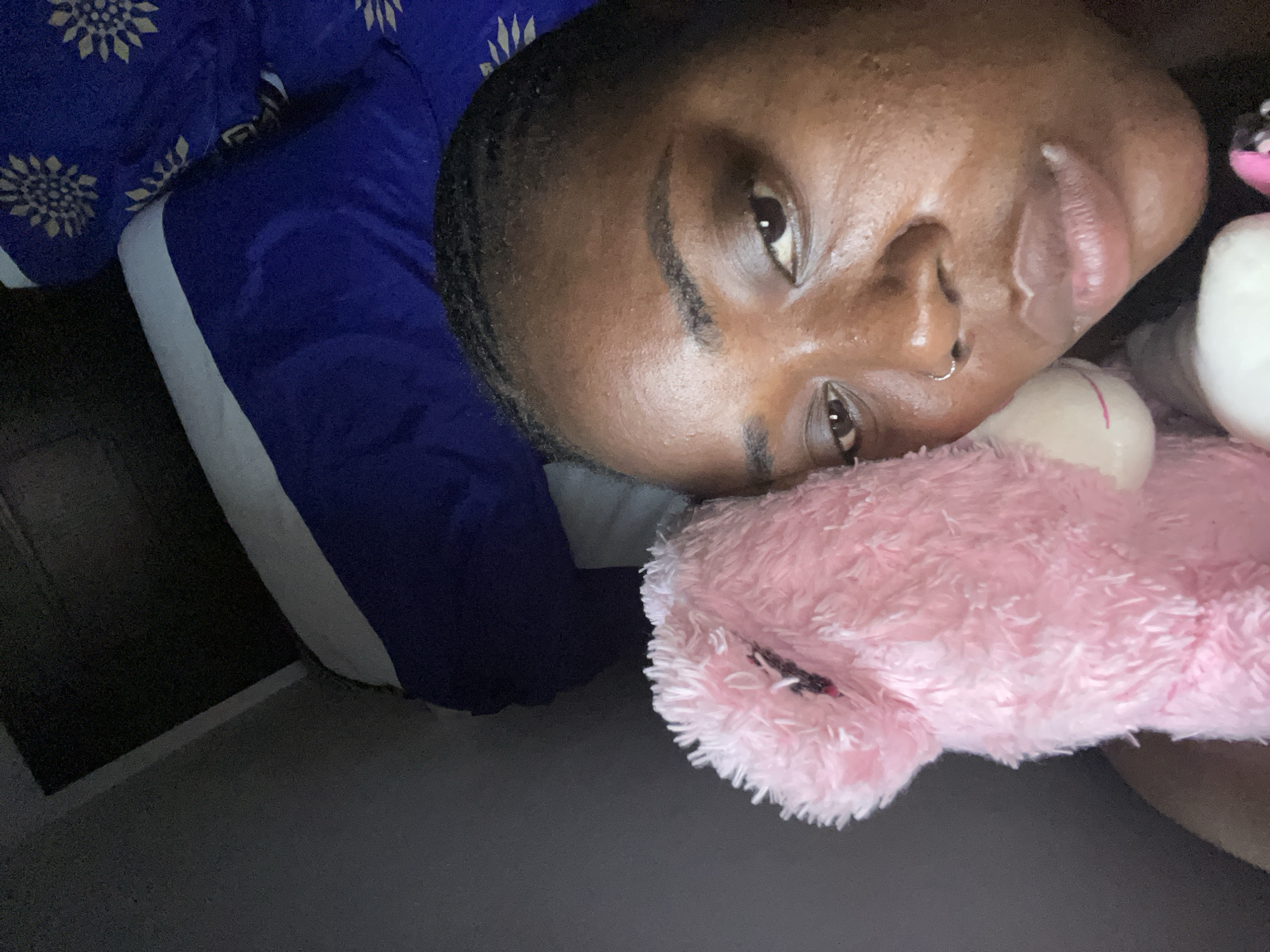 A person with dark skin, short dark hair, and dark eyes is depicted in a close-up, lying down indoors. A silver hoop nose ring adorns their right nostril. They are looking directly at the camera with a neutral, relaxed expression, their lips slightly parted.
The person is resting their head on or near a fluffy pink plush object, possibly a toy or a soft pillow/blanket. A blue fabric with a white stripe is visible, along with another blue fabric featuring a white and gold floral or starburst pattern, suggesting a blanket or bedding. A small portion of a white object, possibly another plush toy, is also seen beside the pink one.
The scene is dimly lit, indicating evening or nighttime, and appears to be an indoor setting, likely a bedroom, in Abuja, Nigeria. No visible text is present in the image.