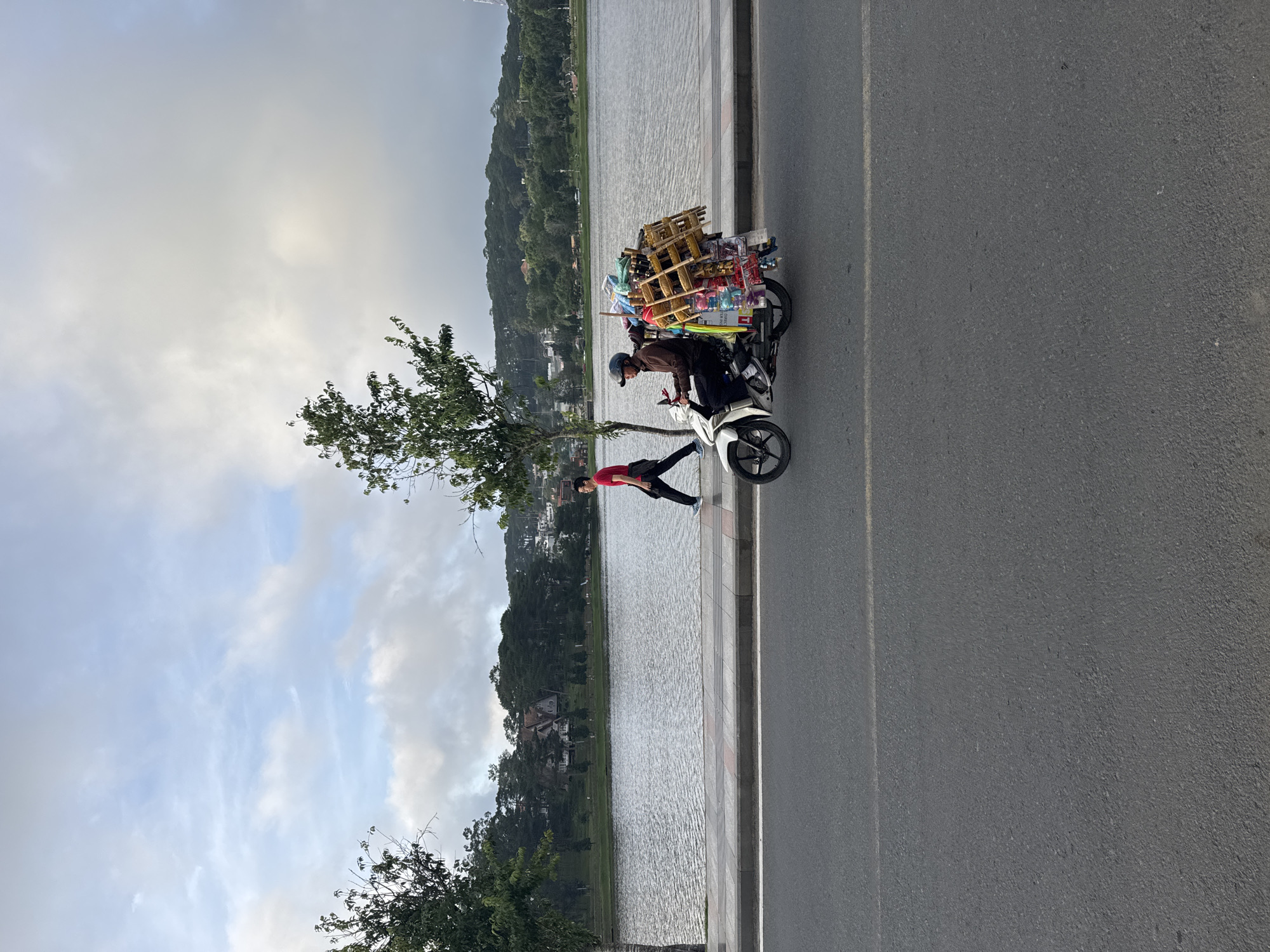 The image displays an outdoor scene in Da Lat, Vietnam, depicting a street and lakefront area, consistent with Xuan Huong Lake. In the foreground, a grey asphalt road runs parallel to a concrete sidewalk, separated by a dark curb. Beyond the sidewalk lies a expansive body of water with a slightly rippled surface, leading to a distant shoreline populated by numerous green trees and several residential buildings nestled among hills. The background features overcast sky with scattered white and grey clouds.
Two male individuals are present, both appearing to move from left to right. On the sidewalk, a man wears a red short-sleeved shirt, dark trousers, and light athletic shoes, captured mid-stride. On the asphalt road, a second man operates a white scooter-style motorcycle. He is dressed in a dark long-sleeved shirt, dark trousers, and a dark helmet. The motorcycle is heavily laden with various goods, including a prominent multi-tiered wooden display rack filled with numerous small, colorful items such as toys, whistles, and other novelty products, suggesting the operator is a mobile vendor. A tree with green foliage stands on the sidewalk between the two individuals.