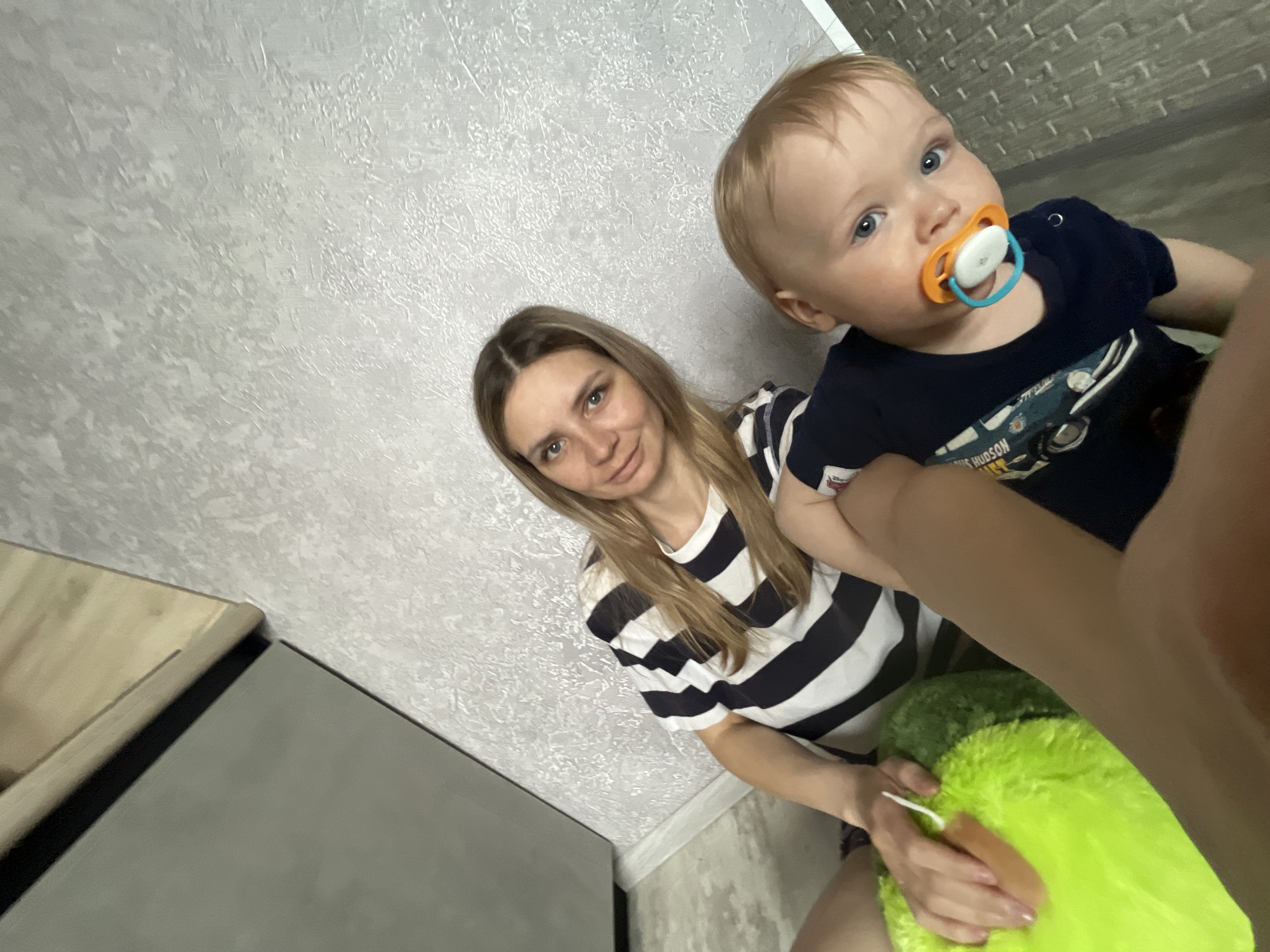 A woman with long blonde hair wearing a black and white striped shirt is sitting next to a baby with blonde hair and a pacifier. The baby is wearing a navy blue shirt. The woman is holding a green stuffed animal. They are in a room with textured gray walls. A gray cabinet is visible on the left. The photograph was taken at an angle from above. This image was taken in the city of Chaikovsky, Russia.