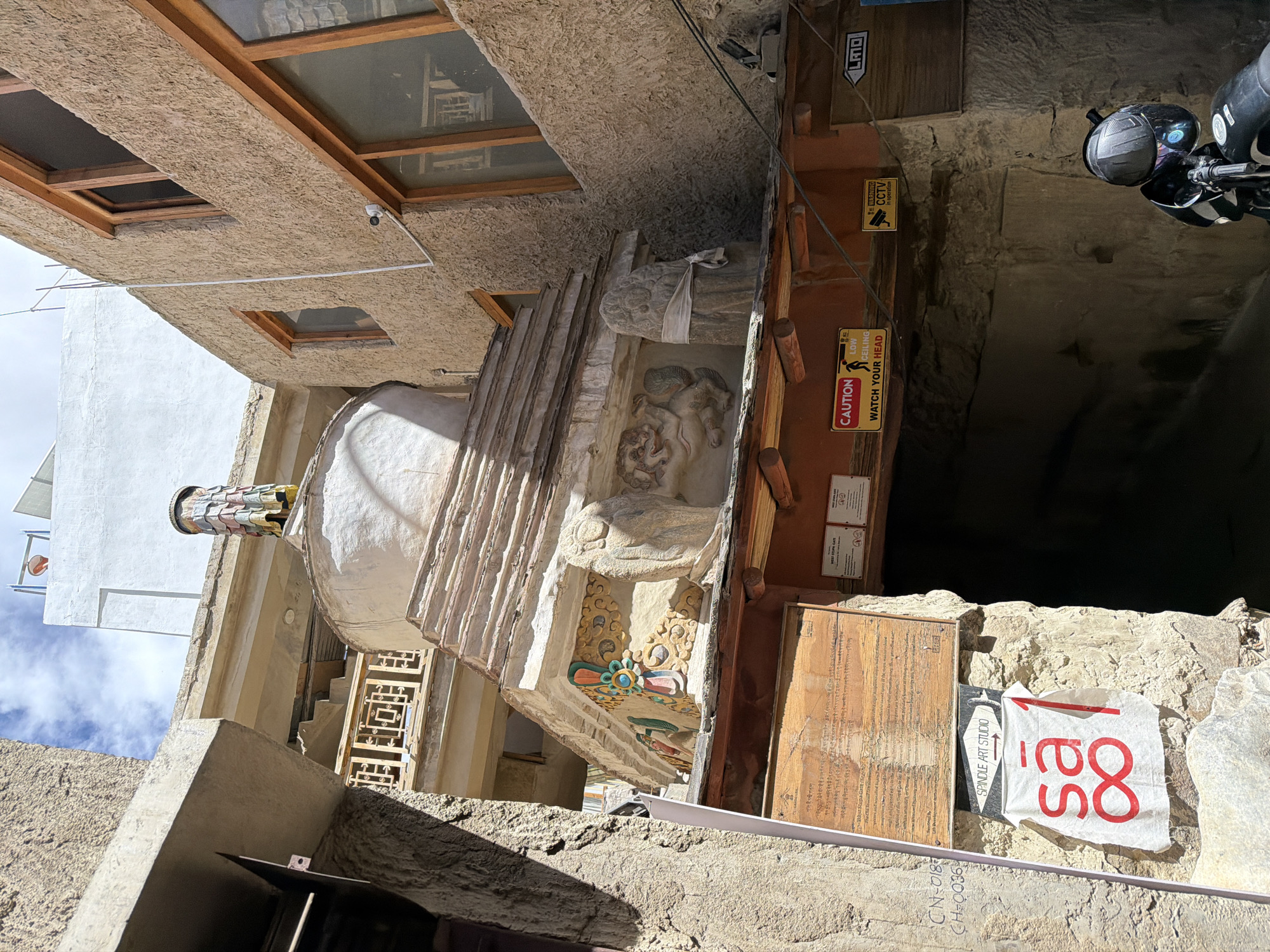 A low-angle outdoor shot captures a narrow alleyway in Leh, India, framed by textured, earth-toned buildings. Dominating the center is a tiered, white stupa, intricately decorated with colorful carvings and what appears to be a fabric or paper topping. To the left of the stupa, a weathered wooden sign with text in an unknown script is affixed to the building. Below this, a white cloth banner with red Arabic-style numerals "sa1" is displayed. To the right of the stupa, the entrance to a dark tunnel or passage is visible. Above the entrance, several warning signs are mounted: one reads "CAUTION LOW CEILING WATCH YOUR HEAD," another features a CCTV camera icon with "WARNING CCTV in operation," and a third sign with an arrow points left and reads "LM". A motorcycle with a black helmet is parked in the lower right corner. The sky above is a mix of blue and white clouds.