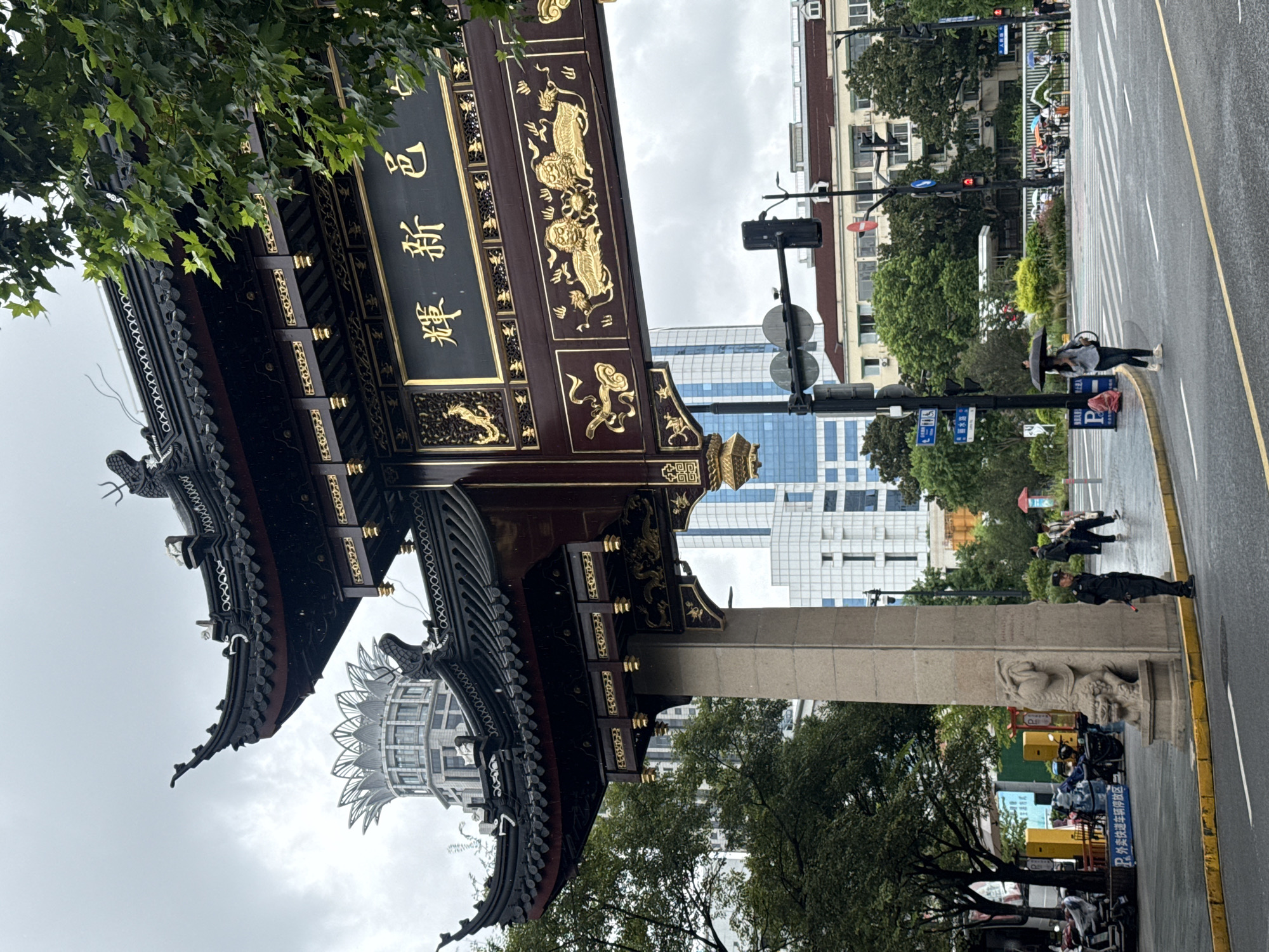 The image depicts a street scene in Shanghai, China, specifically at the entrance to Yu Garden, a historic Chinese garden. In the foreground, a large, ornate Chinese-style gate stands prominently. The gate is dark wood with intricate gold detailing, featuring traditional Chinese motifs such as dragons and lions. The roof is tiered and has upturned eaves. A stone pillar supports the gate, and a carved stone lion statue sits at its base.
Behind the gate, modern Shanghai architecture rises, including tall buildings with glass facades and a distinct, lotus-shaped building in the distance. Trees line the street, and the sky is overcast, suggesting it might be raining, as evidenced by the light streaks appearing to be raindrops.
Several people are visible in the scene. A man in a dark uniform, possibly a security guard, stands near the stone pillar. Other pedestrians are crossing a street with a marked crosswalk, some holding umbrellas. Vehicles, including motorcycles and cars, are present on the road. Traffic lights and street signs are also visible, indicating a busy urban environment. The overall atmosphere is one of a bustling city where traditional and modern elements coexist.