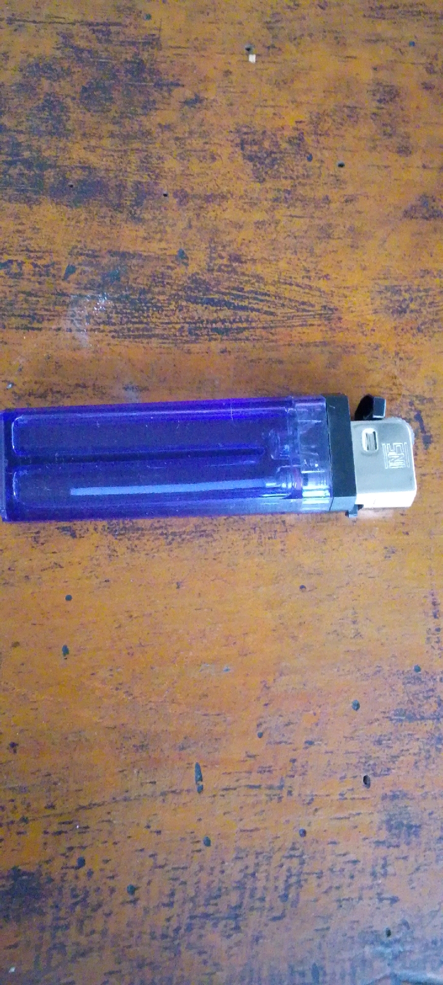The image shows a purple lighter lying horizontally on a wooden surface. The lighter is transparent, allowing a glimpse of its inner workings. The wooden surface has a rustic texture with visible grain and some darker, irregular marks. The lighting suggests an indoor environment. The image does not provide any information about the city of Ngaglik, Indonesia.