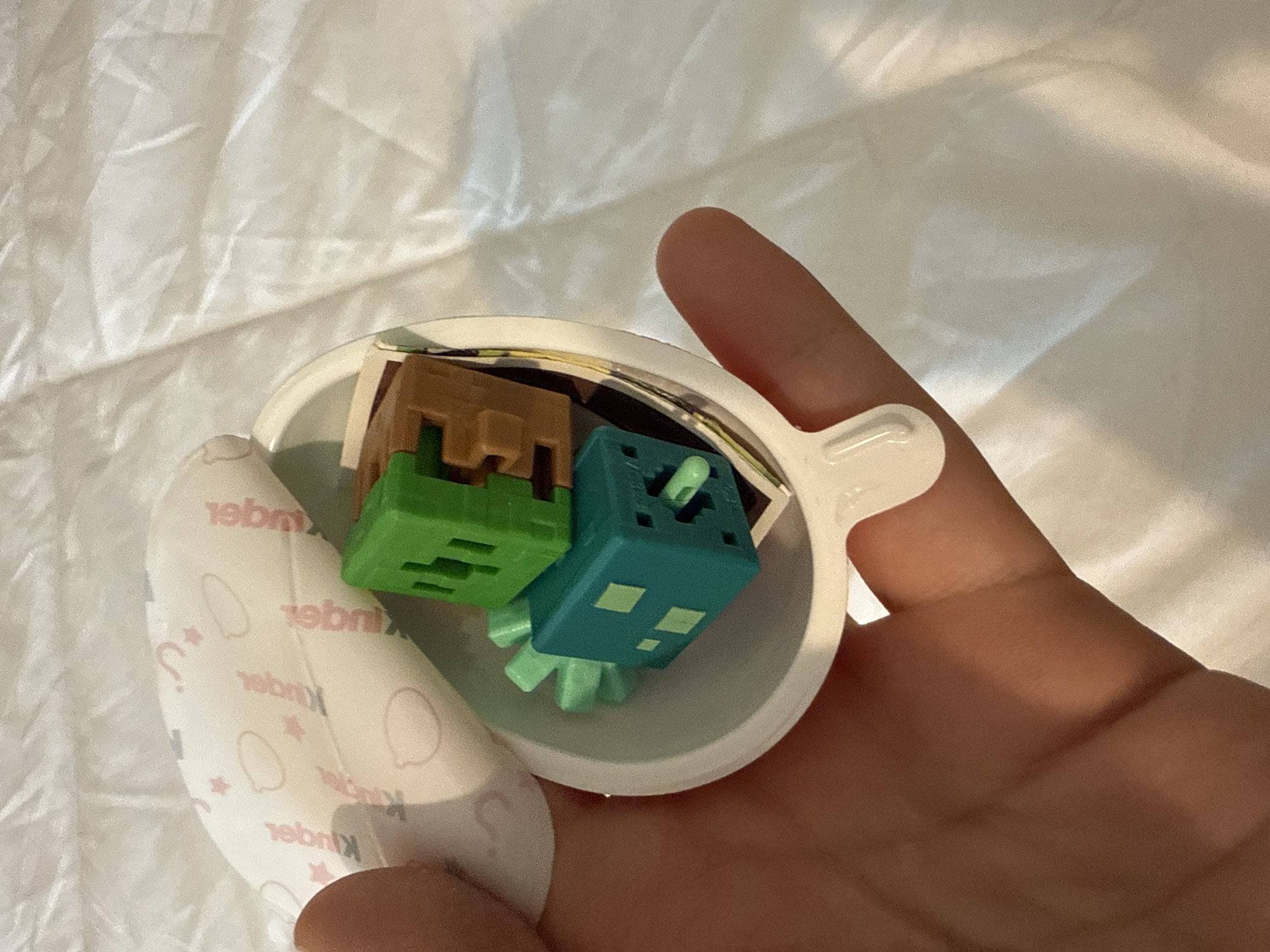 A close-up image shows a person's hand holding an open white plastic capsule from a Kinder Surprise egg. Inside the capsule are two small, block-shaped plastic toys, reminiscent of characters from the video game Minecraft. One toy is brown and features a blocky, angular design. The other toy is teal or blue-green with lighter green accents, also having a cubic form and what appears to be small appendages or legs at its base. Behind the toys, a folded paper insert, likely instructions or a collectible guide, is partially visible.
To the left, a portion of the white and patterned paper wrapper from the Kinder egg is visible, with the word "Kinder" printed multiple times in reverse, along with subtle illustrations of egg shapes and question marks. The background is a soft, light-colored, possibly white or off-white, textured fabric, such as a sheet or blanket, which is slightly wrinkled. The hand has a natural skin tone and appears to be in the process of holding or presenting the revealed contents of the Kinder egg. The lighting suggests an indoor setting, with no specific time of day discernible. No explicit location cues are visible within the image itself.