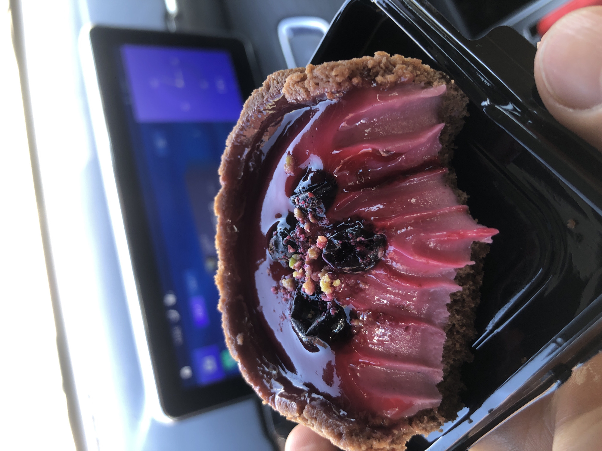 The image presents a close-up view of a partially consumed dessert, identified as a tart or small pie, held by a human hand within an automobile interior. The dessert features a textured, dark brown crust. Its interior consists of a two-layered filling: a vibrant fuchsia-pink, cream-like substance with distinct piped ridging, which is exposed where a bite has been taken from the front-center of the dessert; and an overlying, glossy, dark red-purple glaze. Scattered across the glaze are several dark, shriveled berries and fine, light-colored granular particulates. The dessert rests within a black, ridged plastic container, which is grasped by a left hand, with the thumb and index finger visible on the left side of the frame. The background is out of focus, revealing elements of a vehicle's interior, including a digital infotainment screen displaying a blue user interface in the mid-ground. Above this, a brilliant, overexposed light source suggests bright outdoor conditions viewed through a car window. A blurred, white, indeterminate object is discernible on the far right edge of the background. The specific location within the city of Saint Petersburg, Russia, cannot be ascertained from the visual information provided in this image.
