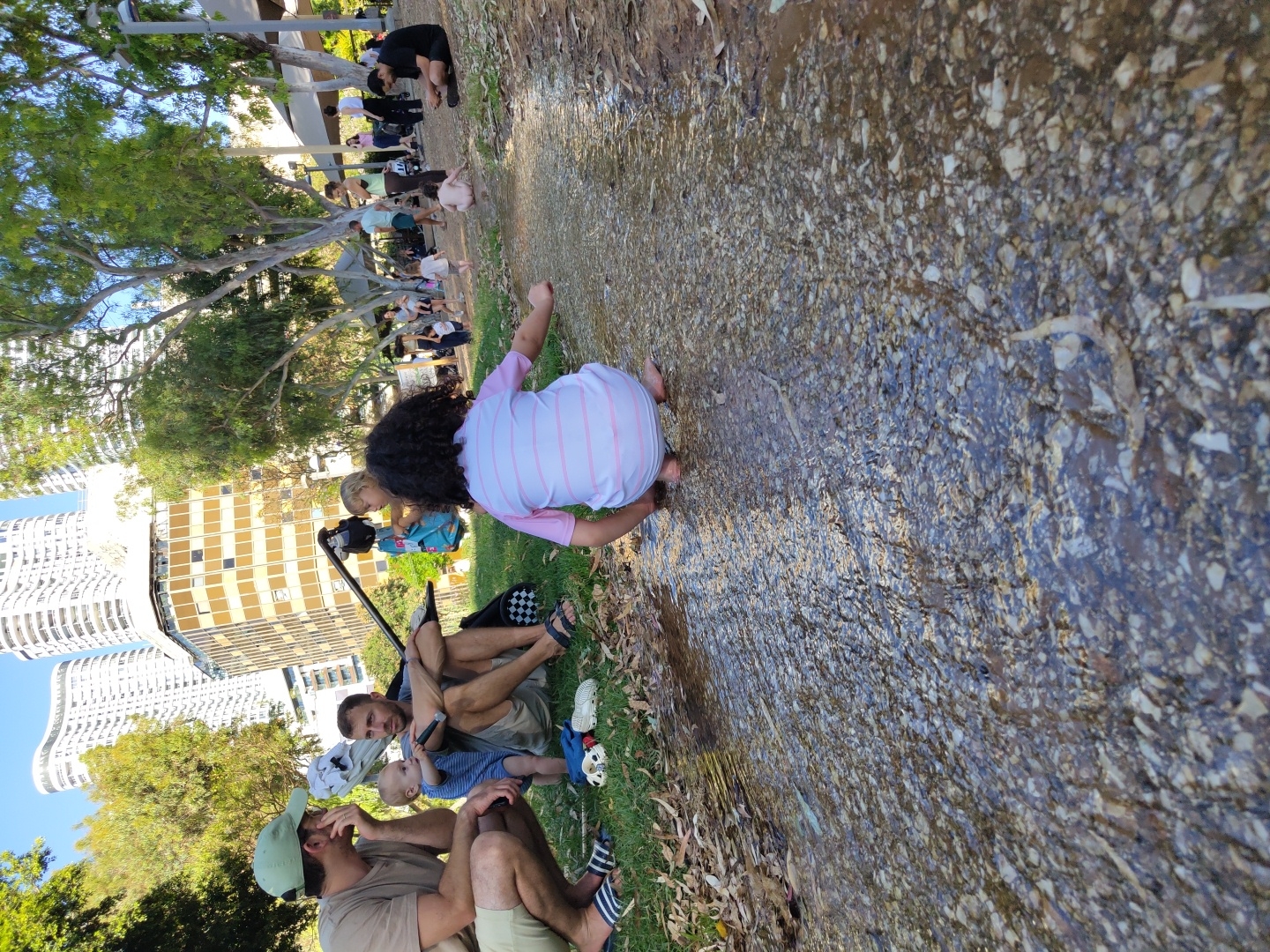 The image captures a scene in a public park in Tel Aviv, Israel, characterized by lush greenery, tall trees, and modern urban architecture in the background. In the foreground, a shallow stream of water flows over a rocky bed. A young child, facing away from the camera, is crouched at the edge of the stream, their back to the viewer. The child is wearing a pink and white striped outfit. To the left of the child, two adults and a baby are seated on the grass. One adult, wearing a green cap and a light-colored t-shirt and shorts, is squatting. Beside them, another adult, wearing a blue and white striped shirt and shorts, is sitting. A baby in a blue and white striped outfit is held by the sitting adult. The background reveals a bustling park environment with other people, some sitting, some standing, and children playing. Tall, white high-rise buildings dominate the skyline, indicative of Tel Aviv's urban landscape. The presence of a filming rig with a camera and boom microphone suggests that a video or film might be in production. The overall atmosphere is relaxed and family-oriented, set against the backdrop of a modern city.