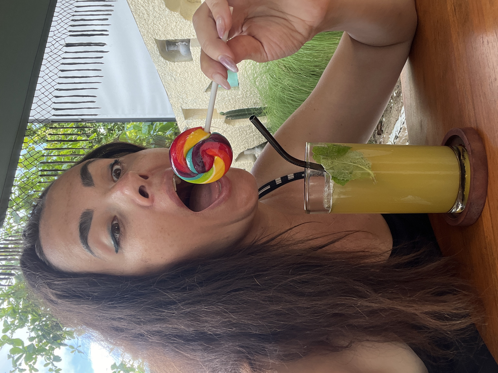 A woman is shown from the chest up, holding a rainbow lollipop to her open mouth and looking at the camera. She has dark brown hair and is wearing a black top. In front of her is a glass of yellow liquid with mint leaves and ice, and a dark straw. The background shows greenery and a building with a textured wall, suggesting an outdoor setting. The overall impression is one of a casual, possibly vacation-related moment. The location is identified as Kuta Selatan, Indonesia.