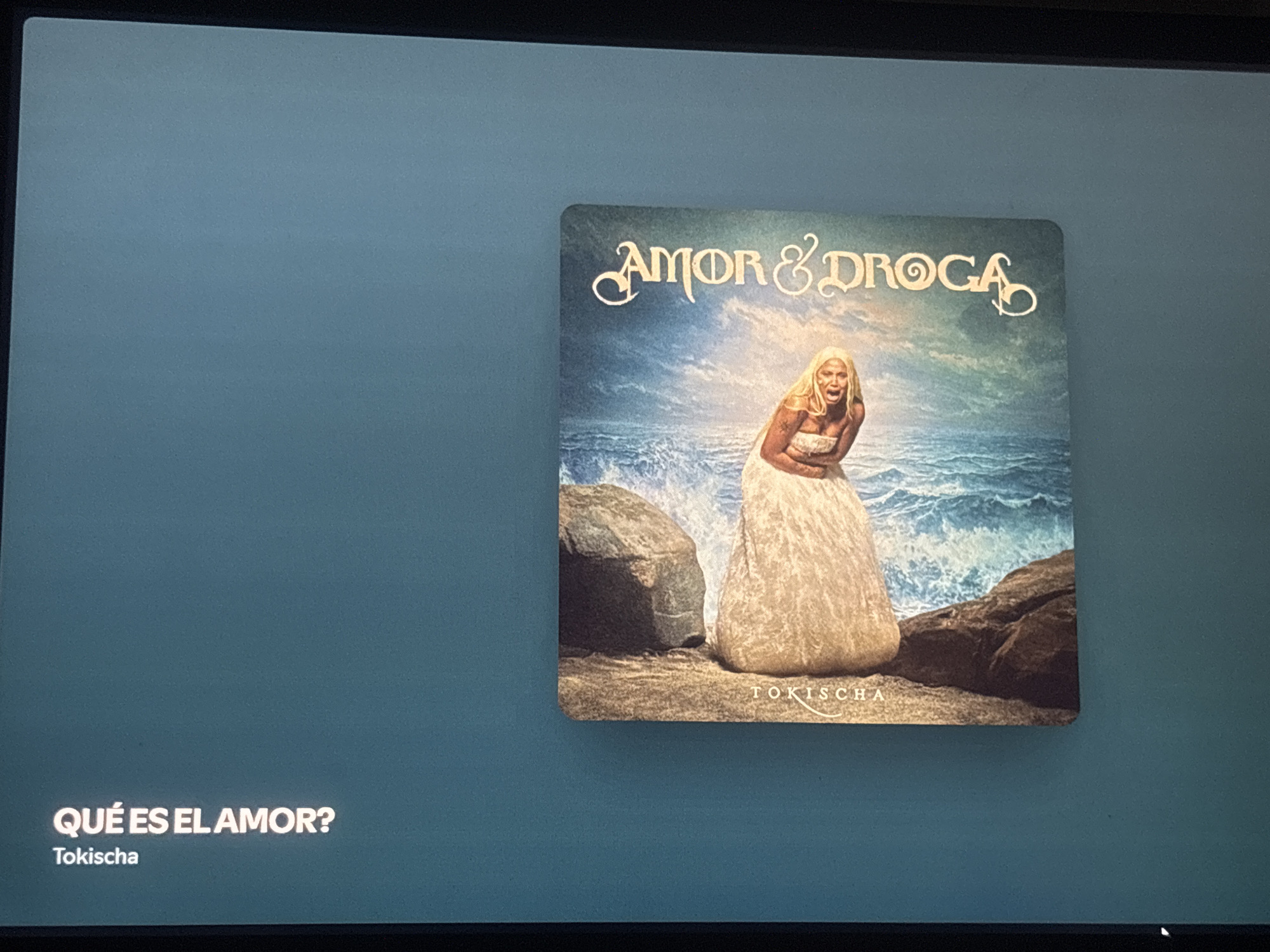 The image shows a screen displaying the cover art and information for the song "Qué Es El Amor?" by Tokischa. The album cover, titled "Amor & Droga," features a woman in a white dress standing on a rocky beach with the ocean in the background. The woman has blonde hair and appears to be in distress, with her mouth open as if screaming. The song title and artist's name are displayed in white text on the lower left side of the screen. There is no indication of Buenos Aires, Argentina, in the provided image.