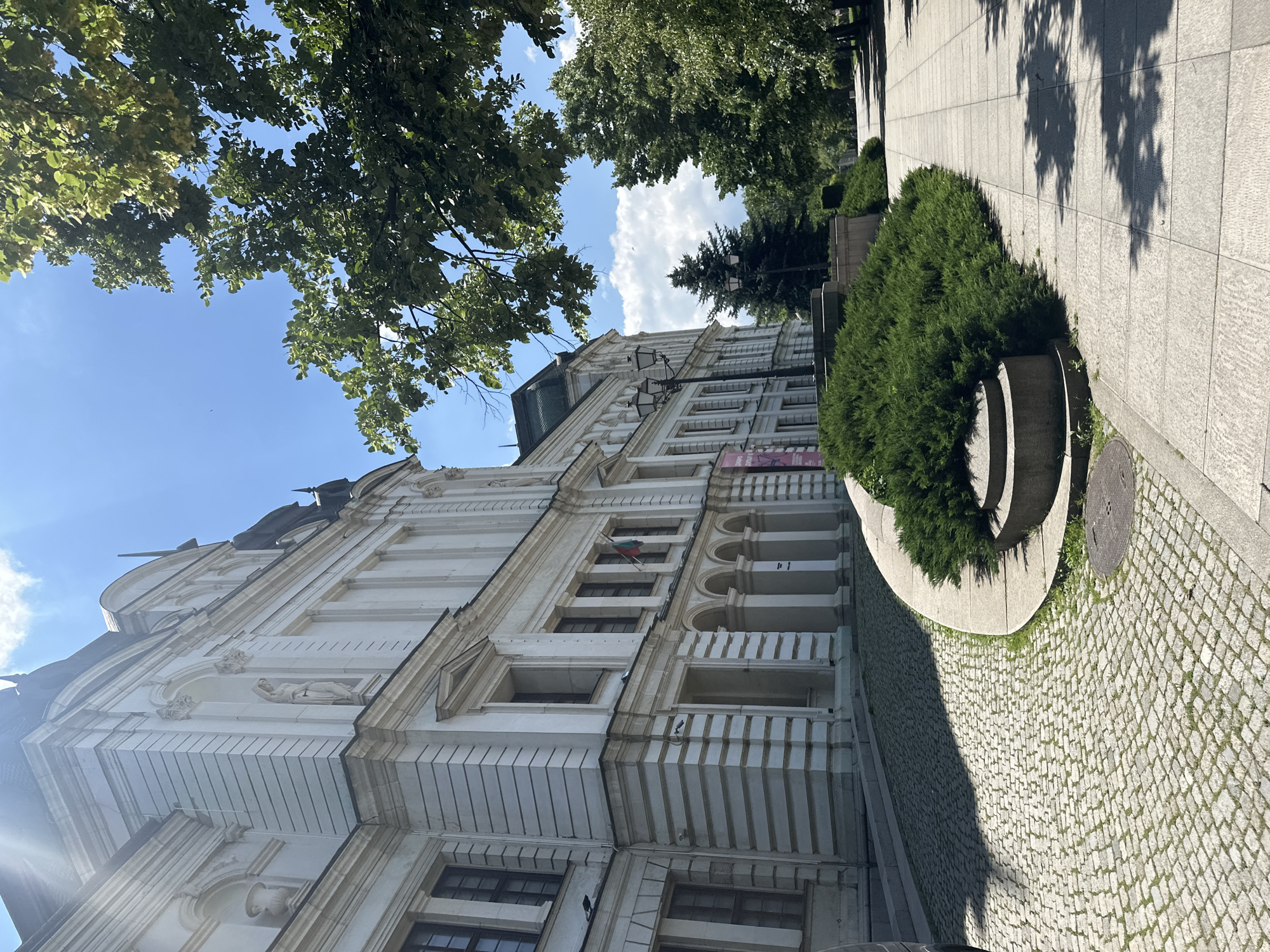 The image shows the Ivan Vazov National Theatre in Sofia, Bulgaria, on a bright, sunny day. The facade of the theatre is white with neoclassical architectural details, including columns, arches, and statues. A Bulgarian flag is visible hanging from the building. In the foreground, a cobblestone pathway curves around a large, manicured hedge. To the right, a wide paved sidewalk runs alongside a tree-lined street, with the shadows of the leaves dappling the ground. The sky is a clear blue with some scattered white clouds. No people are visible in the scene.