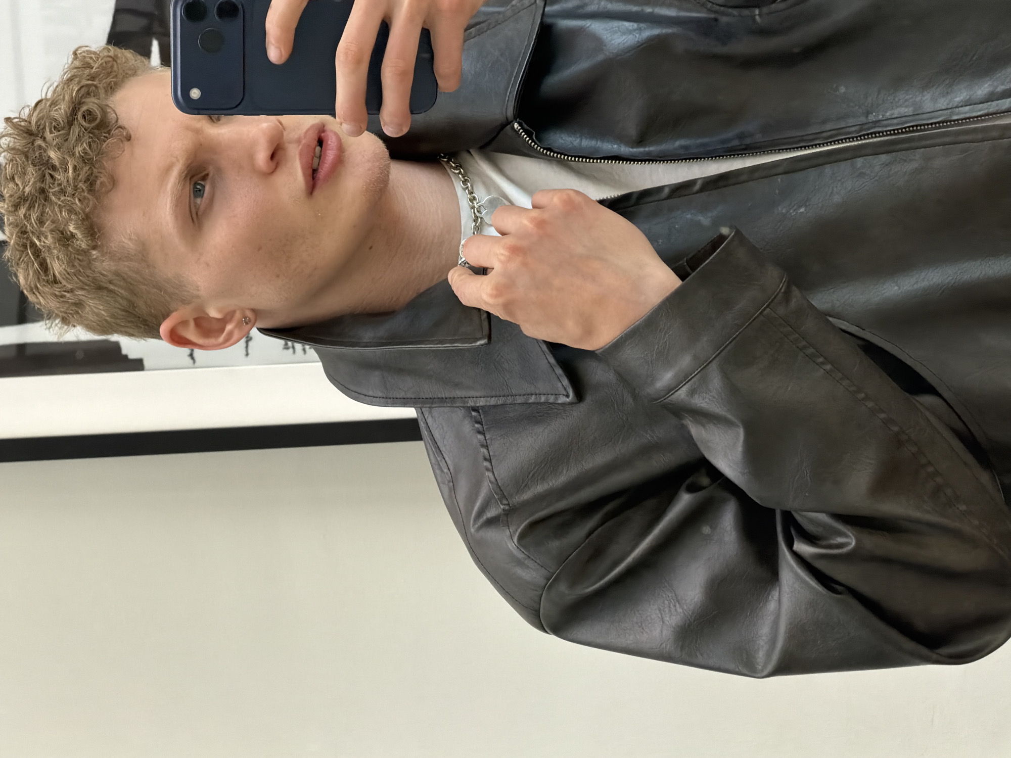 A young man is captured in a selfie taken indoors in London, United Kingdom. He has fair skin, short, curly blonde or light brown hair, and blue eyes. He is wearing a dark, likely black or very dark brown, leather or faux leather jacket over a white t-shirt. A silver chain necklace with a heart-shaped pendant is visible around his neck, which he is touching with his left hand. A small silver stud earring adorns his left ear.
He holds a dark blue smartphone up with his right hand, angled to take the photo, and his gaze is directed towards the phone's camera. His lips are slightly parted, and his expression is neutral or mildly pensive. The background consists of a plain light-colored wall with a dark, horizontal line running across it, suggesting a simple indoor setting. The lighting appears to be ambient indoor light. No text is visible in the image.