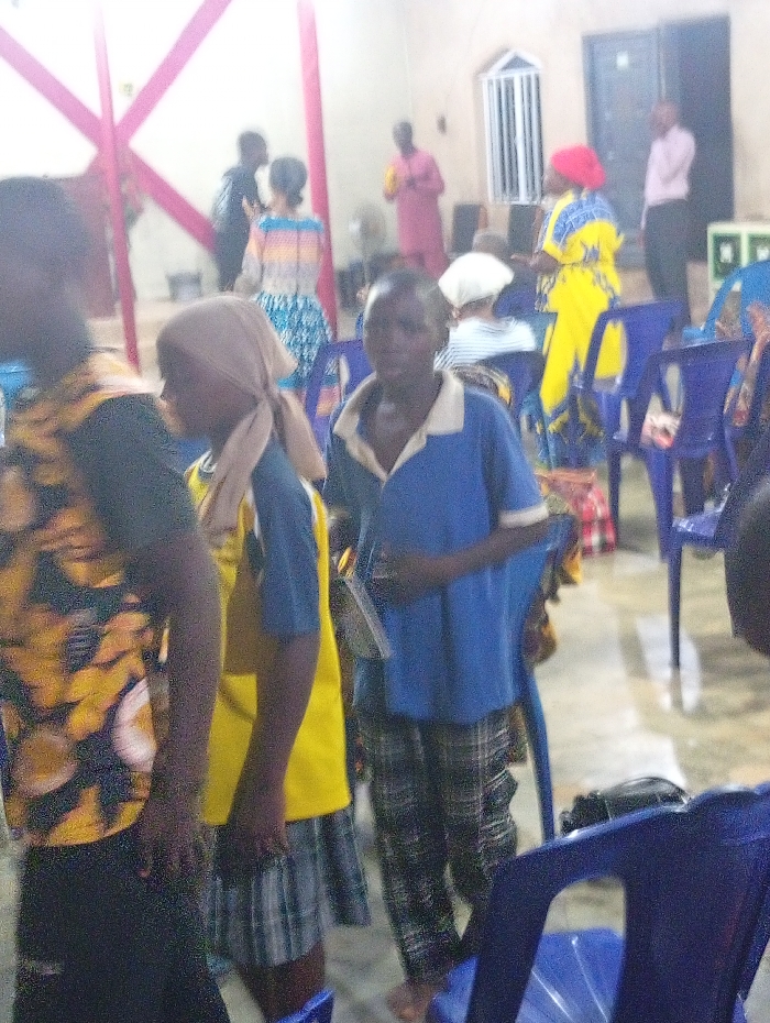 A group of people are indoors, likely at a church or gathering place. Several individuals are in the foreground, including two children, one in a blue shirt and checkered pants. A third child is wearing a yellow and blue shirt, with a head covering. Further back, several adults are standing, some wearing colorful clothing. Chairs are scattered around, and the lighting seems dim. The scene appears somewhat blurred and grainy.