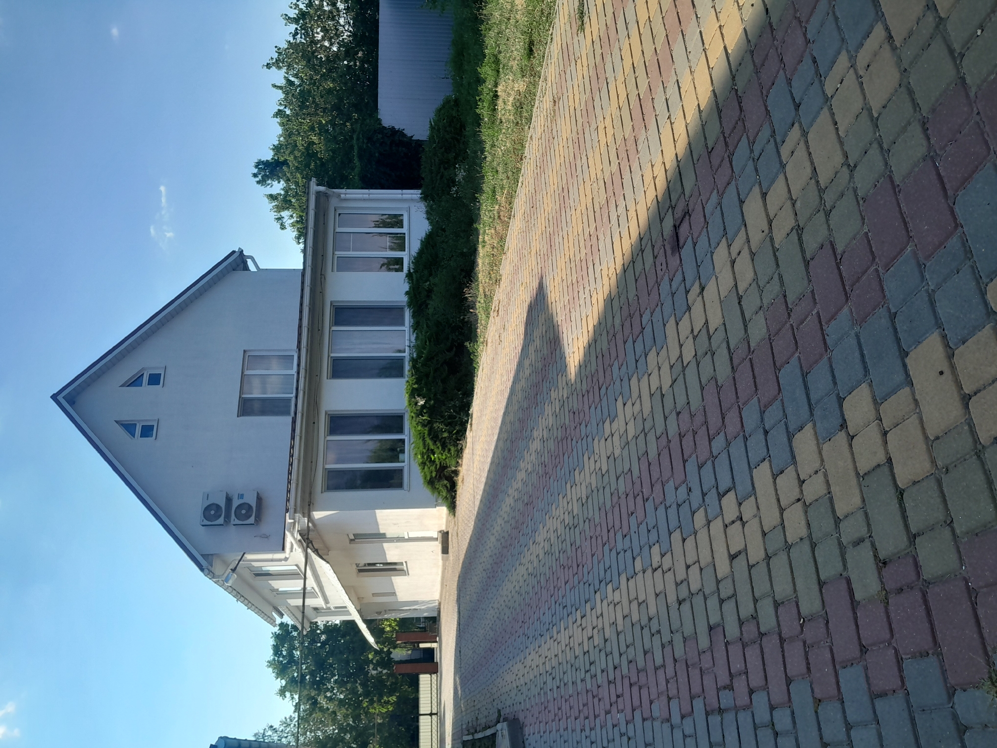 The image shows a two-story white building with an attic, situated in a paved driveway. The building has several windows, including two large ones on the first floor and several smaller ones on the second floor and attic. Two air conditioning units are mounted on the exterior wall of the second floor. The driveway is paved with multi-colored rectangular bricks. Green trees and bushes surround the building, and a metal fence is visible in the background. The sky is clear with some scattered clouds. The location is the city of Illinka, Ukraine.