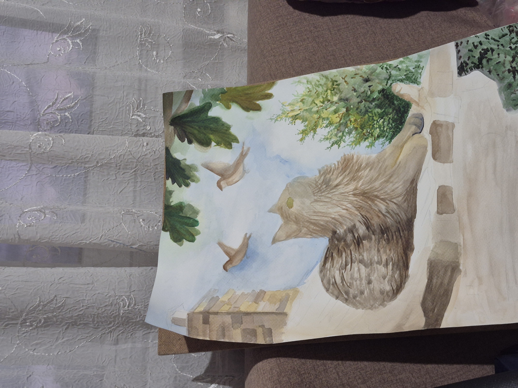 The image displays a watercolor painting of a cat lying on a brick ledge, with two birds flying in the sky above and a tree with green leaves. The painting is propped up against a couch, and a textured white curtain with embroidered patterns is visible in the background. The setting appears to be indoors, with the painting placed in front of a window with the curtain. No specific location in the city of Popovo, Bulgaria is discernible from the image itself. The painting is unfinished, with some areas showing only pencil sketches. The cat is depicted in shades of brown and gray, with its head turned slightly to the left and its eyes closed. The birds are small and brown, with their wings outstretched. The tree is rendered in various shades of green, with detailed leaves. The brick ledge is painted in earthy tones, with some bricks appearing lighter than others. The overall scene is serene and captures a moment of quiet observation by the cat.