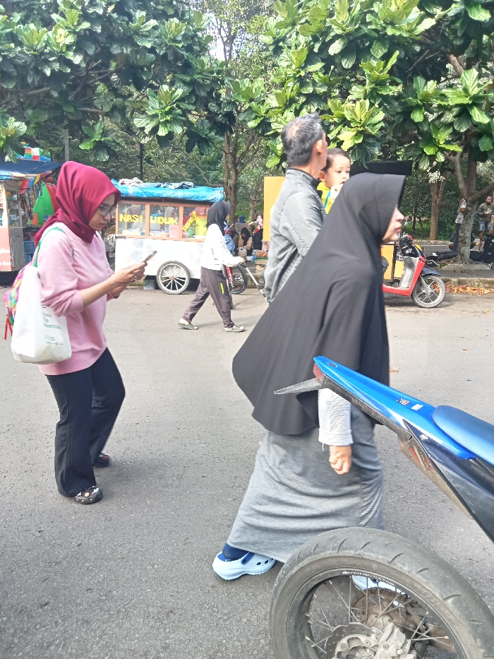 The image captures a lively outdoor scene in Singaparna, Indonesia, bustling with pedestrians. In the foreground on the left, a woman wearing a maroon hijab, a light pink long-sleeved top, dark pants, and patterned clogs is engrossed in her smartphone. She carries a white tote bag with a vibrant strap.
Further into the scene on the right, a man with graying hair, dressed in a gray striped long-sleeved shirt, carries a small child whose head and yellow top are visible. He walks beside a woman wearing a long black hijab that drapes over her torso, a gray long skirt, and white Crocs-style shoes with blue socks.
In the mid-ground on the left, behind the woman on her phone, another person in a white top and dark hijab pushes a bicycle. A blue food cart, featuring the prominent text "NASI UDUK," stands nearby, indicating a local street food vendor. A parked red scooter is visible on the right side of the mid-ground, and the rear wheel and blue fender of a motorcycle are in the extreme foreground on the right.
The setting is an asphalt path or road, lined with numerous large trees featuring broad, green leaves, suggesting a park, market area, or public thoroughfare. Several other people can be seen in the background, some near stalls. The scene is brightly lit by natural daylight, indicating it is daytime. The overall impression is one of everyday activity in a casual, public space.