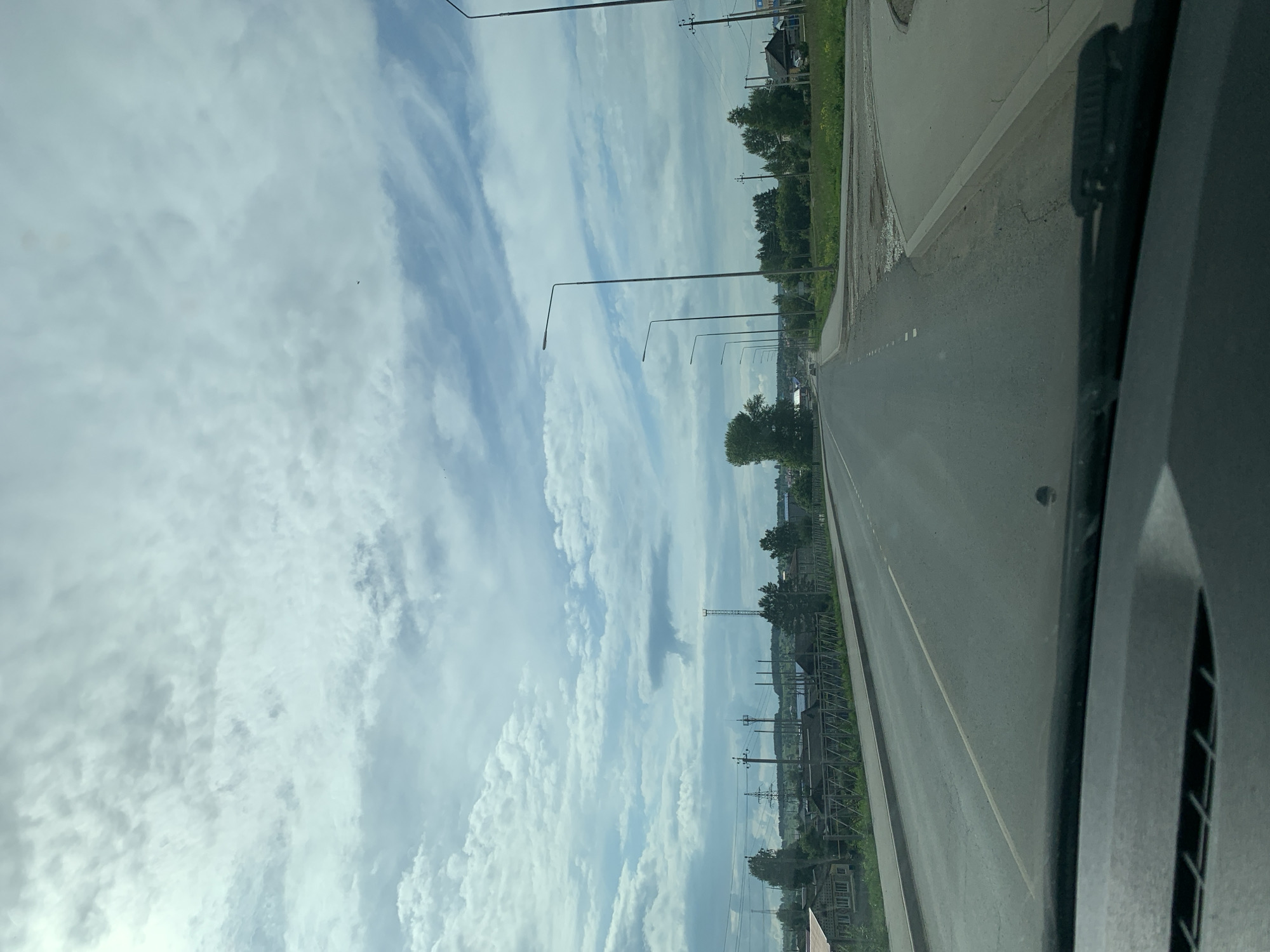 The image captures a view from a vehicle driving on a paved road in the city of Orda, Russia. The road stretches ahead, with a clear lane marking in the center. On the left side of the road, there are houses and a wooden fence, indicating a residential area. Trees and utility poles are visible along the roadside. The right side of the road features a sidewalk and a grassy area with more trees and buildings in the distance. Streetlights line the road, suggesting it is an urban or suburban environment. The sky above is partly cloudy, with a mix of white and gray clouds against a light blue backdrop. The overall scene depicts a typical day in a Russian city, with infrastructure and natural elements coexisting.
