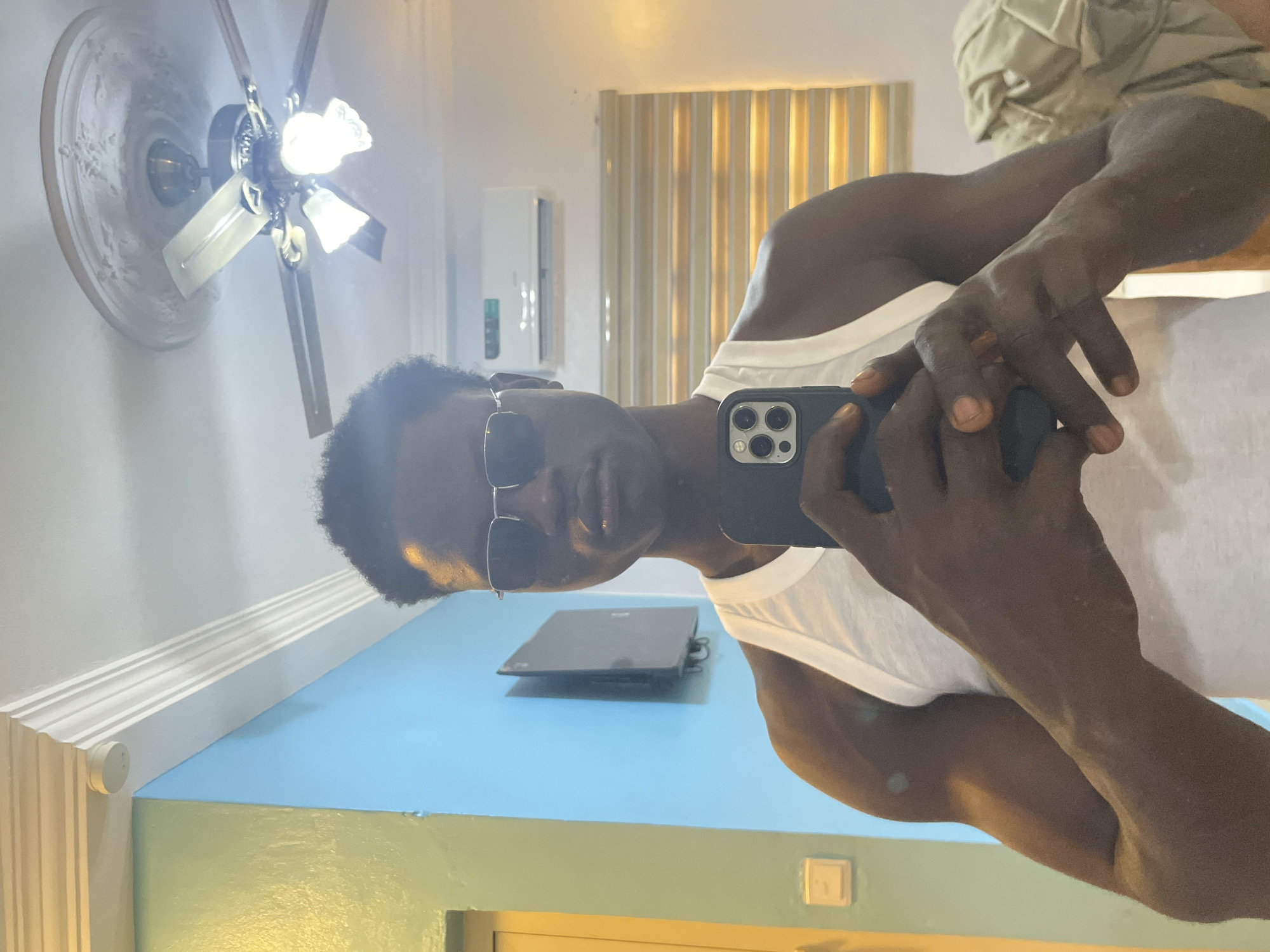 A man wearing a white tank top and sunglasses is taking a selfie in what appears to be a room. He is holding a phone with a triple-lens camera. In the background, there is a ceiling fan, a beige curtain, and what seems to be an air conditioning unit mounted on the wall. The lower portion of the wall is painted blue, and the lower part of the wall is yellow. Based on the appearance of the room and assuming the location of Badagry, Nigeria, the scene could be a room in a residential home, a guest house, or a small hotel.