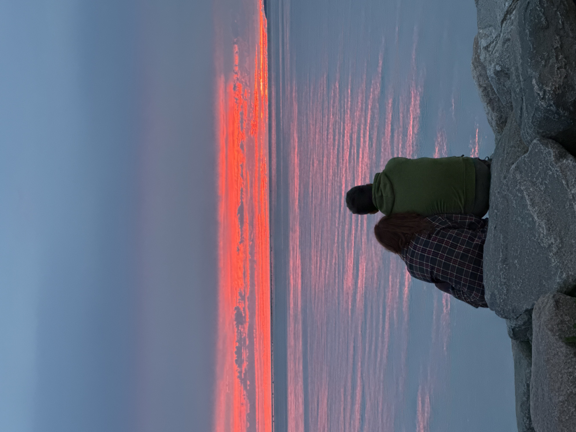 A couple is sitting on large rocks by the water, looking out at a vibrant sunset over the Baltic Sea. The sky is a dramatic gradient of deep blue at the top, transitioning to fiery orange and red where the sun is setting, with wispy clouds catching the light. The water reflects the colors of the sky, creating a serene and colorful scene. The couple is seen from behind, with one person resting their head on the other's shoulder, suggesting intimacy and shared enjoyment of the view. The rocks they are sitting on appear to be part of a breakwater or a shoreline path. The overall atmosphere is peaceful and romantic, capturing a moment of quiet contemplation against a stunning natural backdrop. This could be a scene from Tallinn's coastline, perhaps near Pirita or the Old City harbor, where such sunsets are common.
